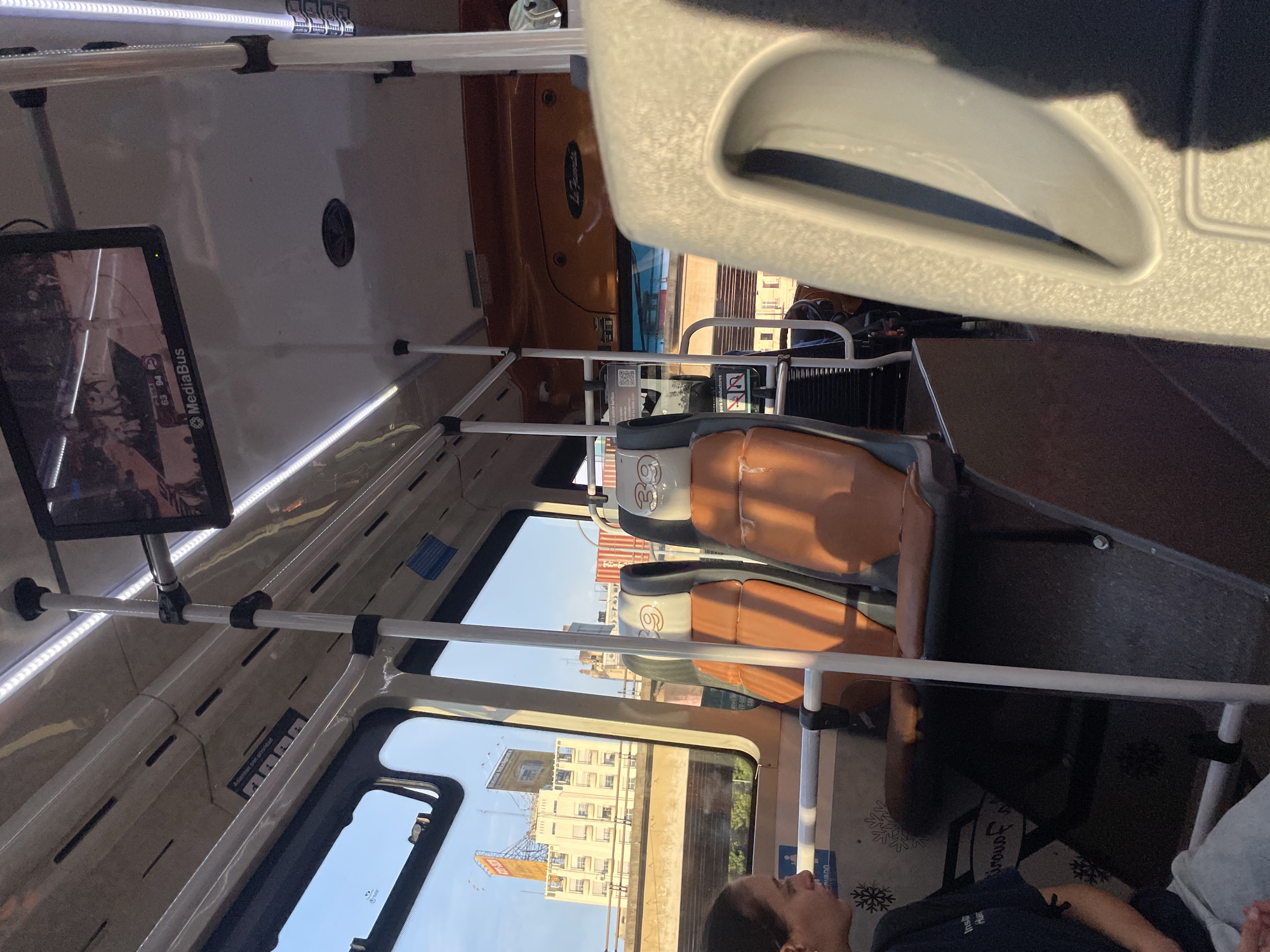 The image depicts the interior of a bus in Buenos Aires, Argentina. A passenger is visible at the bottom of the frame, looking out the window. Outside, buildings are visible, including one with a billboard of a "365" logo. Inside the bus, there are seats and a screen showing "MediaBus". Overhead handrails are visible. The overall impression is that the photo was taken from inside a bus while traveling through Buenos Aires.