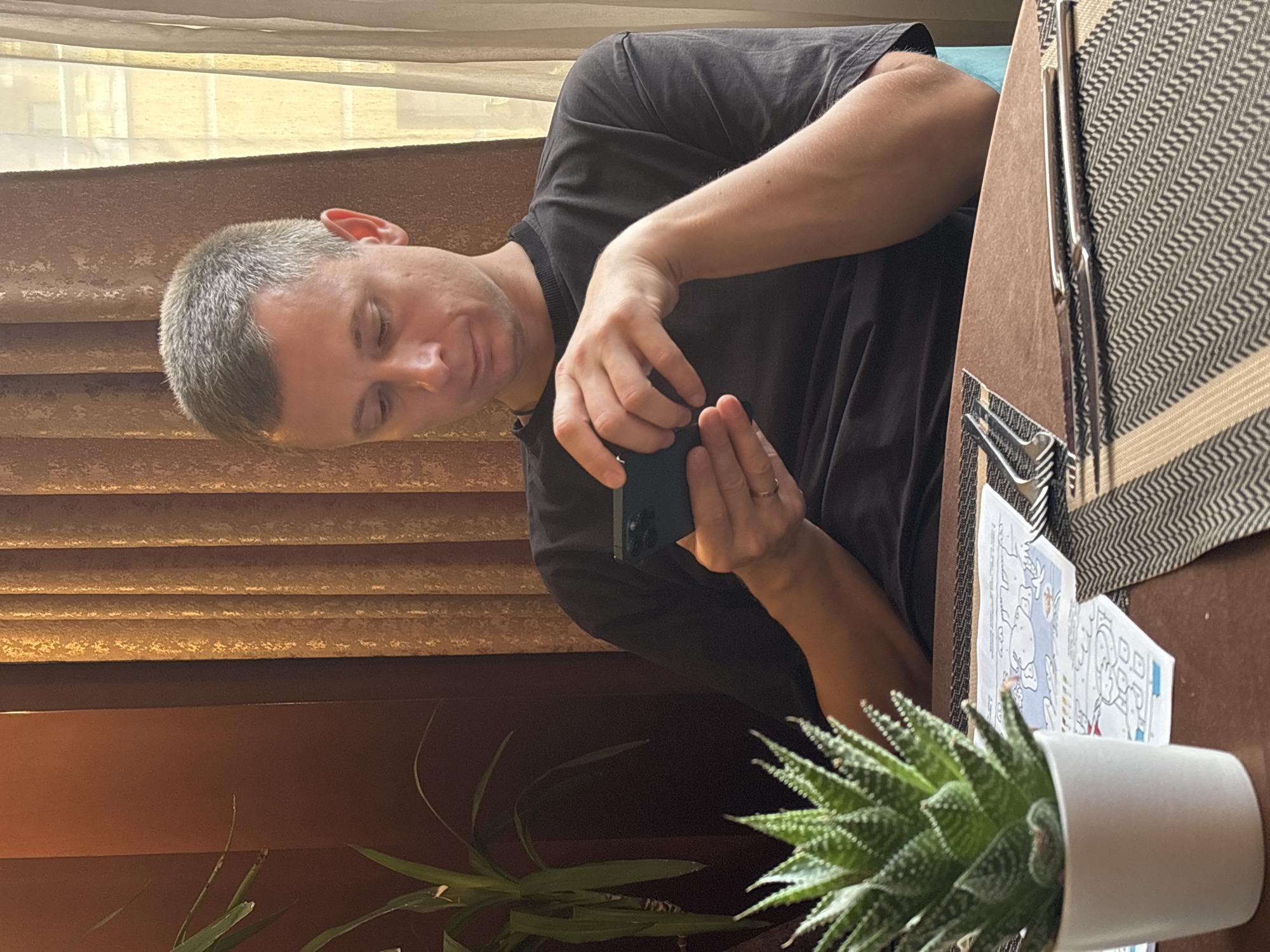 A man with short, graying hair is seated at a table, looking down at his smartphone. He is wearing a dark t-shirt. His left arm rests on the table, and his right hand holds the phone. A small succulent plant in a white pot is to his left, and a placemat with cutlery and a children's placemat lies on the table in front of him. In the background, there are vertical blinds or curtains. The setting appears to be an indoor dining area, possibly a cafe or restaurant, with warm lighting. The image does not provide any specific clues about the location being in the city of Рождествено, Russia.