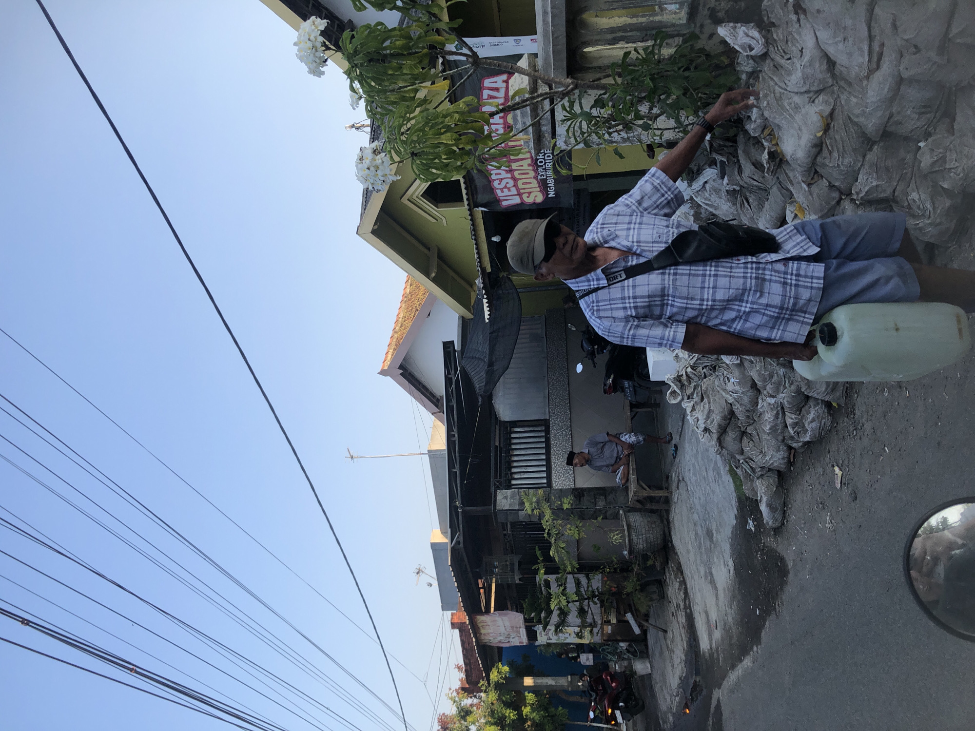 In Kalidawir, Indonesia, a man wearing a plaid shirt, shorts, a baseball cap, and sunglasses walks on a paved street. He carries a large, pale green plastic container with a black lid in his right hand. A black sling bag is worn across his chest. To his right is a large pile of grey, lumpy sacks, possibly sandbags. Behind the man are houses with colorful facades and tiled roofs, some with awnings. A tree with white flowers is visible in front of one of the houses, adorned with a black banner that reads "VESPA SIDOARJO EXPLORERS NGABURIDE". A second man, wearing a grey shirt and checkered shorts, sits on a wooden bench in front of a house on the left. The sky is clear and blue, with numerous power lines crisscrossing overhead. Various motorcycles and potted plants are scattered throughout the scene. The overall impression is of a residential street scene in a tropical climate.