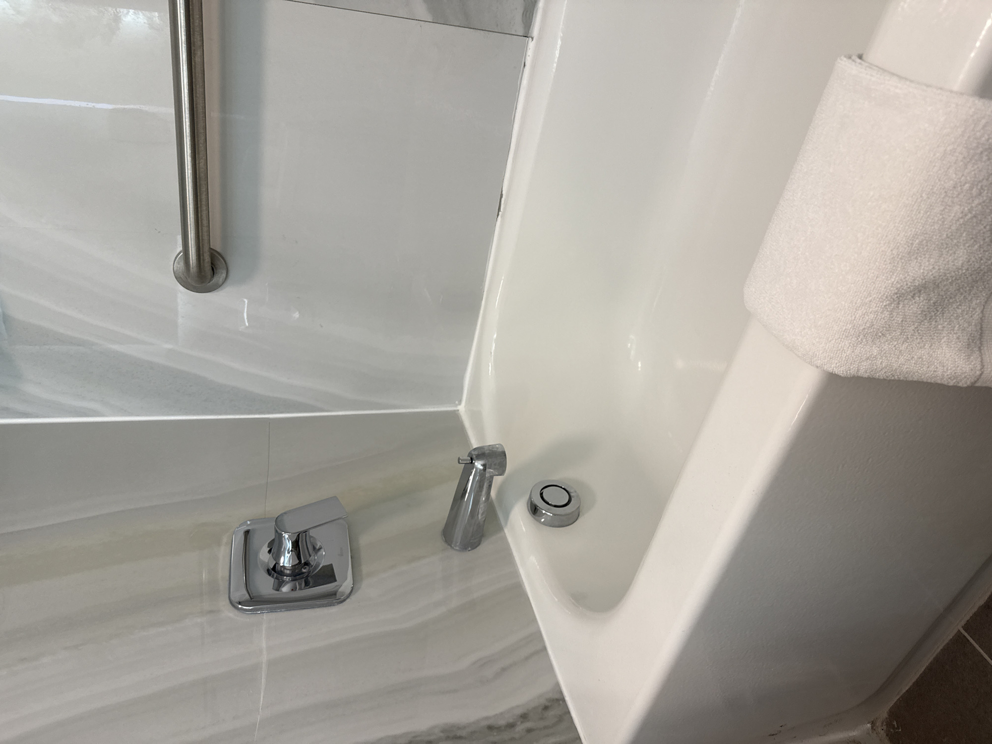 The image shows a close-up view of a bathtub faucet and shower area. The bathtub is white with a chrome faucet set installed on the adjacent surface. A silver grab bar is mounted on the wall next to the bathtub. A white towel is hung over the side of the tub. The countertop and wall are covered in a light gray marble-like material. The location appears to be a bathroom in an indoor setting, possibly a hotel or residence in San Antonio, United States of America, as per the instructions. There are no people visible in the image.