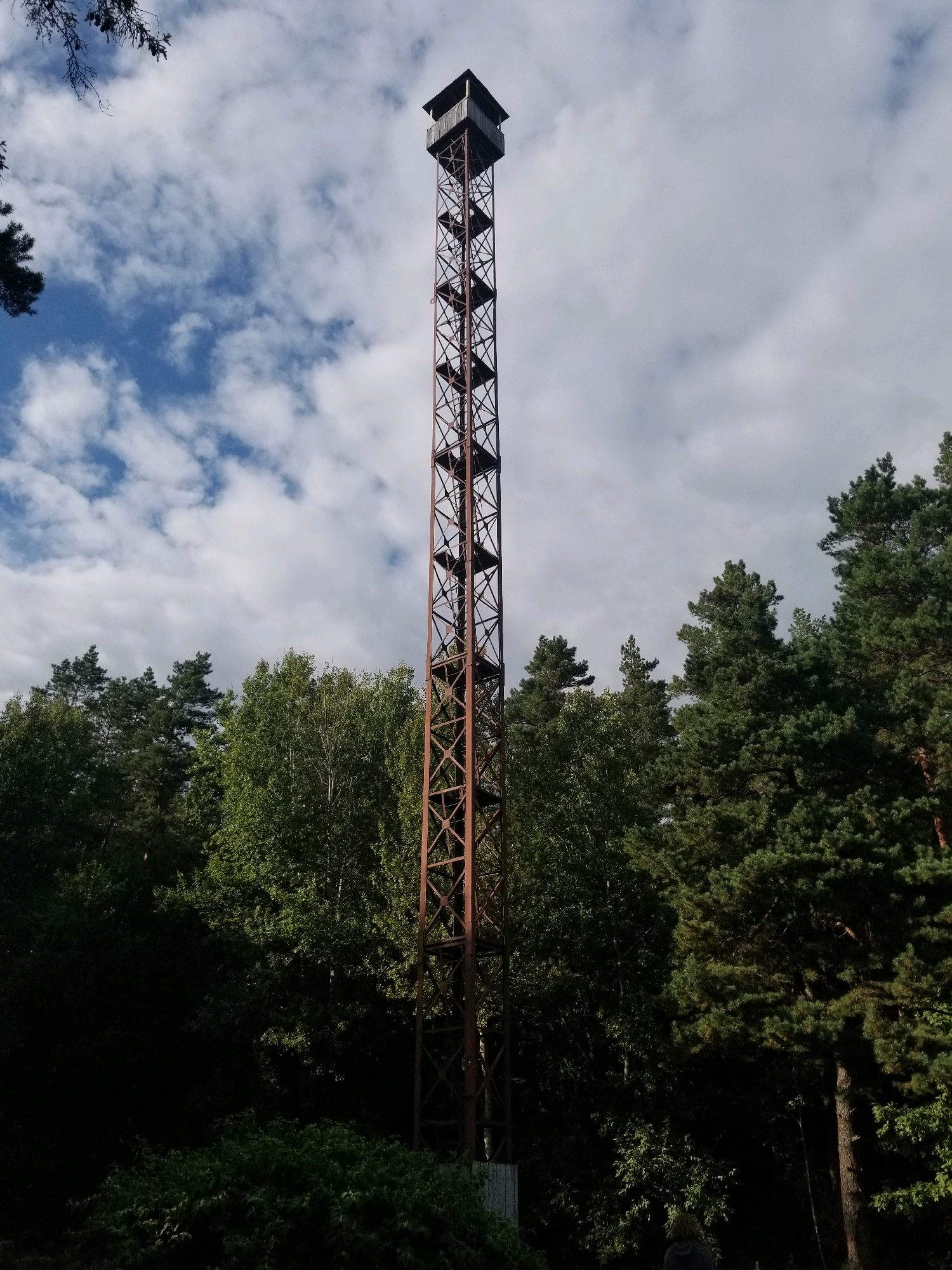 An outdoor, low-angle shot captures a tall, slender, rust-colored metal observation tower against a partly cloudy sky with blue patches. The tower has a lattice-like structure with visible rungs for climbing and a small, enclosed cabin at the very top. Dense, green trees, primarily pine and deciduous varieties, surround the base of the tower, obscuring the lower portion of its structure and the ground. The overall impression is one of a remote, possibly wooded, location. The image's metadata indicates it was taken in Бараўлянскі сельскі Савет, Belarus. There is a person visible at the bottom center of the frame, facing away from the camera, wearing a grey hooded jacket.