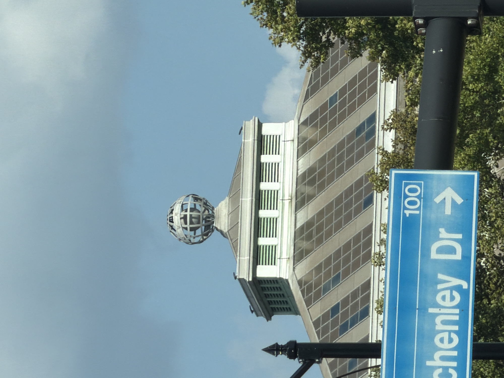 The image shows a building with a geodesic dome structure on its roof, possibly part of the Duquesne Light Company building in Pittsburgh, Pennsylvania. The building has a sloped roof covered in solar panels. In the foreground, a blue street sign reads "100" and "chenley Dr" with a right-pointing arrow. Trees are visible behind the sign. The sky is partly cloudy.