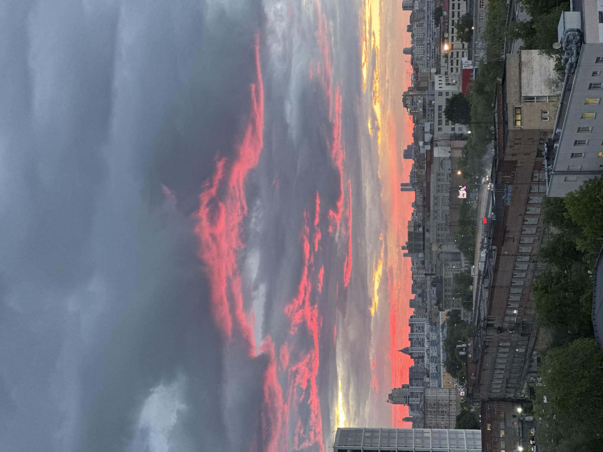 The image shows a cityscape at sunset, likely Moscow, Russia. Tall buildings are visible along the skyline, illuminated by the setting sun which casts a red and orange glow on the horizon. The sky above is filled with grey and pink clouds, suggesting a dramatic weather pattern. Below the skyline, are buildings and trees. A road runs through the buildings. There are very few signs of people.