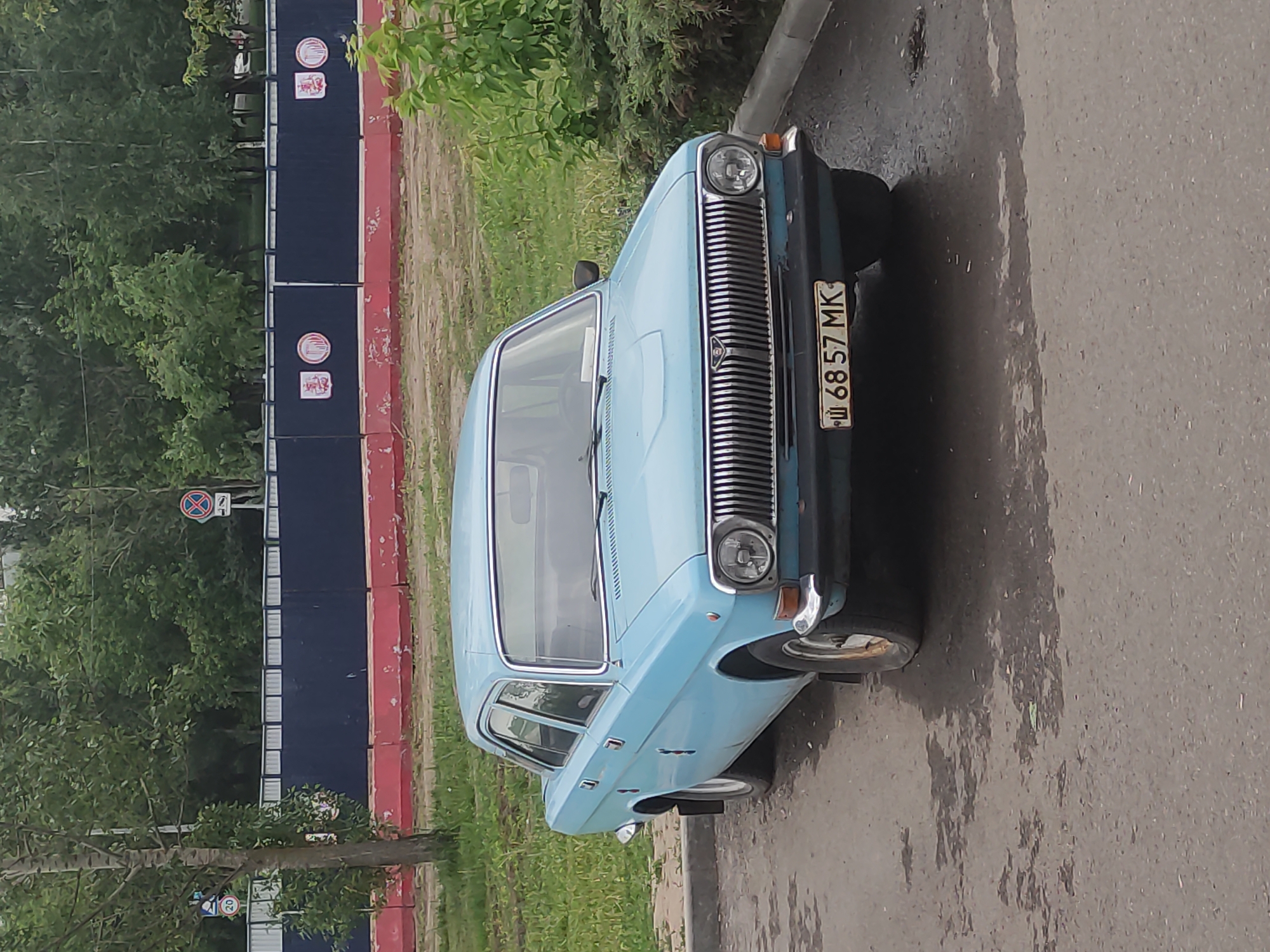 The image depicts a light blue GAZ-24 Volga parked on a street. The license plate number is Ш 68 57 MK, indicating a possible Ukrainian origin. The car appears to be an older model and is located near a blue and red fence, along with some greenery and trees. Based on the presence of the Ukrainian license plate, it is unlikely the location is Moscow, Russia.