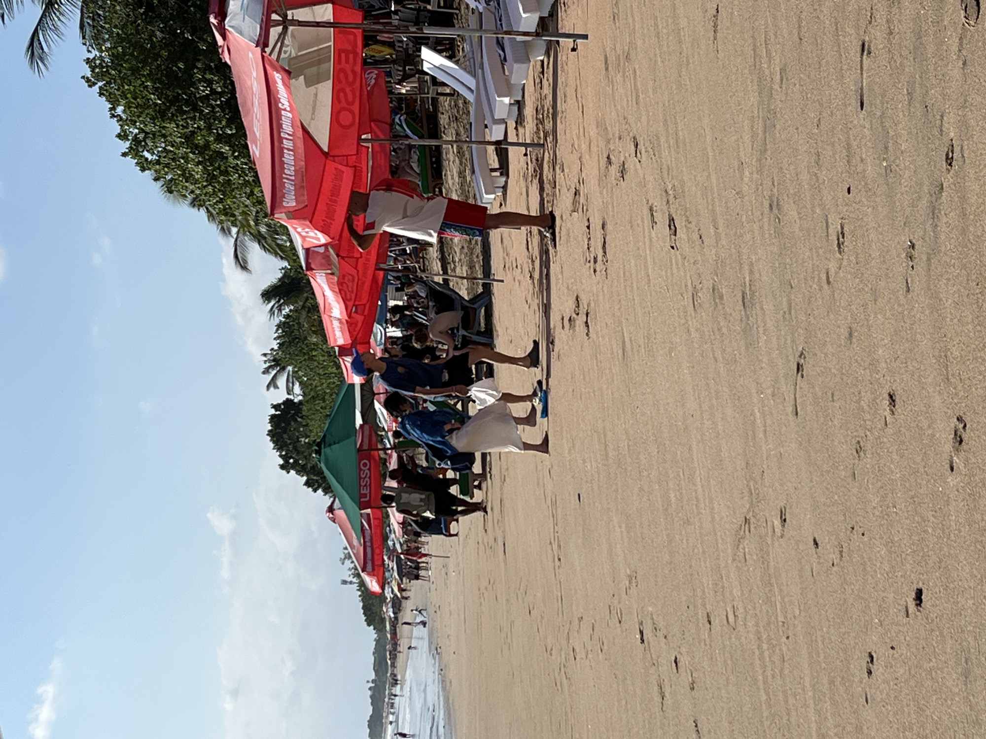 This image depicts a sunny day at Kuta Beach in Bali, Indonesia. The foreground is dominated by a sandy beach with numerous footprints. In the middle ground, a line of red and green umbrellas are set up, with several people interacting beneath them. One person in a blue shirt and white skirt appears to be talking to another individual in a dark shirt. Further back, more beach umbrellas, lounge chairs, and people are visible along the shoreline. The background shows a line of palm trees and a hazy sky. A sign on one of the red umbrellas reads "Global Leader in Piping Solutions".