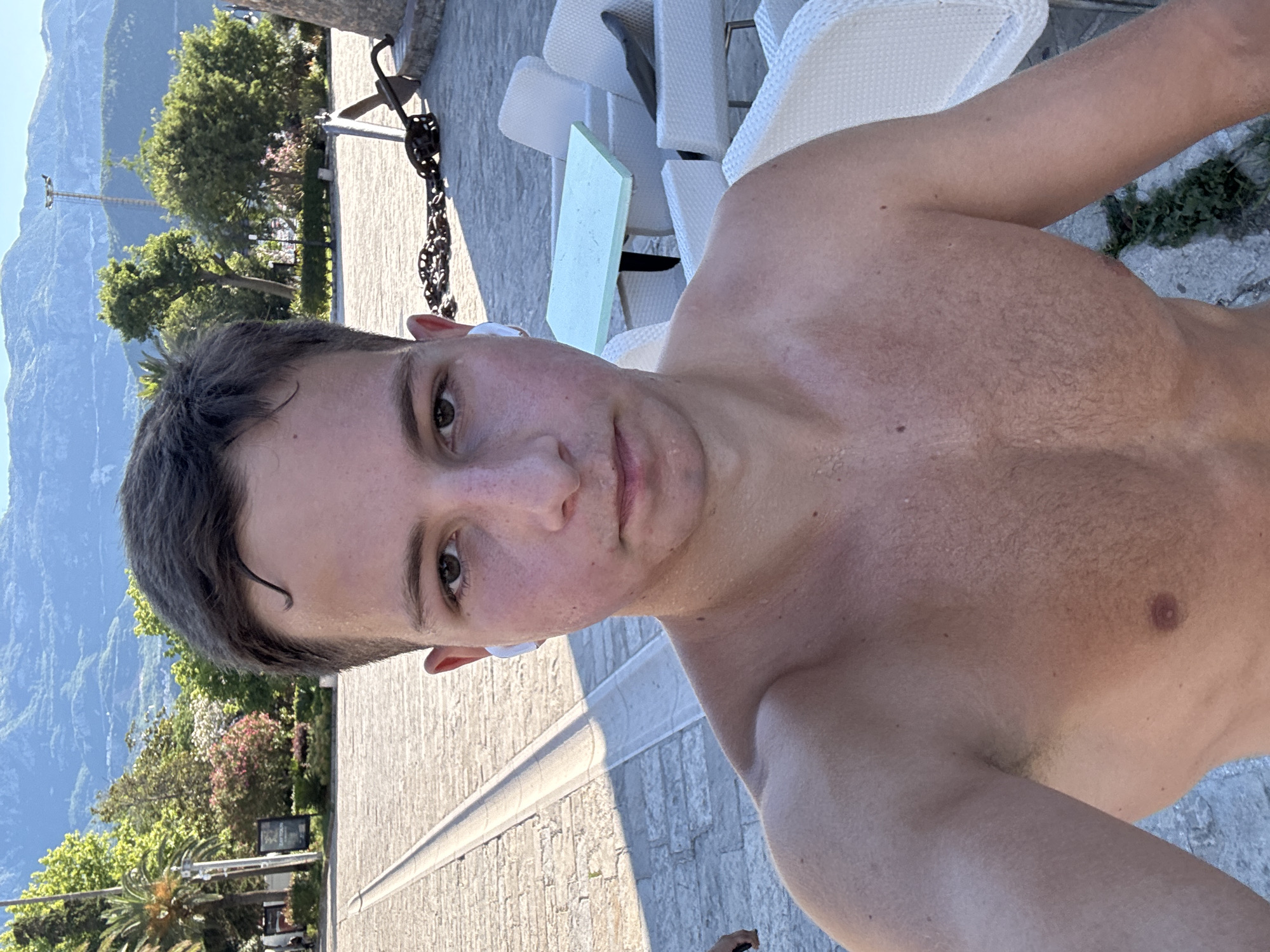 A shirtless young man with short brown hair, wearing white wireless earbuds, takes a selfie outdoors. He has fair skin with some redness and sweat, and sparse chest hair. Behind him, a paved area with a light-colored walkway is visible, with a stone wall on the right. In the background, there are trees, white outdoor chairs and tables, and a mountainous landscape under a clear blue sky. An anchor and chains are positioned near the stone wall. The overall setting appears to be a coastal area in Budva, Montenegro, likely a promenade or public space.