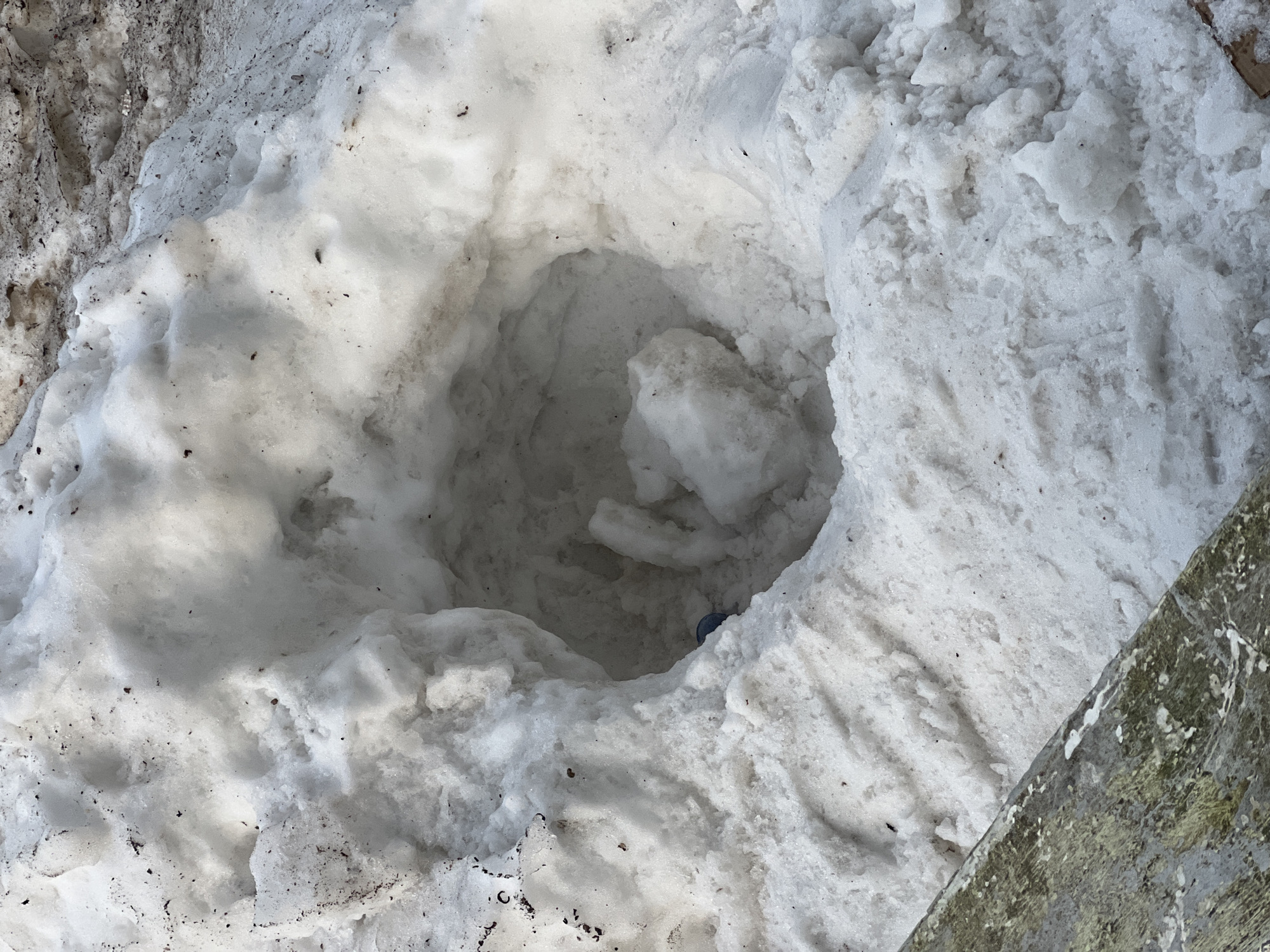 The image shows a hole in the snow. There is some trash at the bottom of the hole, potentially a bottle cap. The surrounding snow is dirty. Part of a wooden plank is in the bottom right corner. The location in Chaikovsky, Russia is unknown.