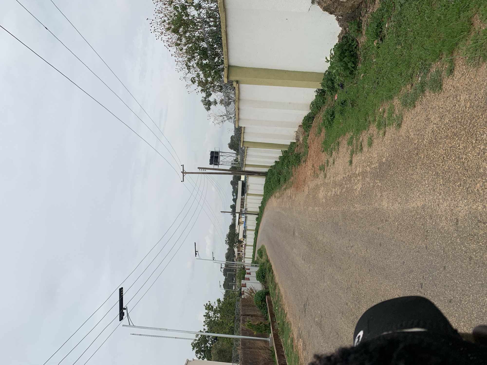 The image depicts a street scene in Barikin Jirga Summa, Nigeria. The view is along a paved road that curves slightly to the right. A tall, white wall with green accents runs along the right side of the road, with vegetation growing on top. Power lines run along the left side of the road, supported by utility poles with streetlights. A water tower is visible in the distance. The sky is overcast. A portion of a person's head, wearing a black cap, is visible at the bottom of the frame.