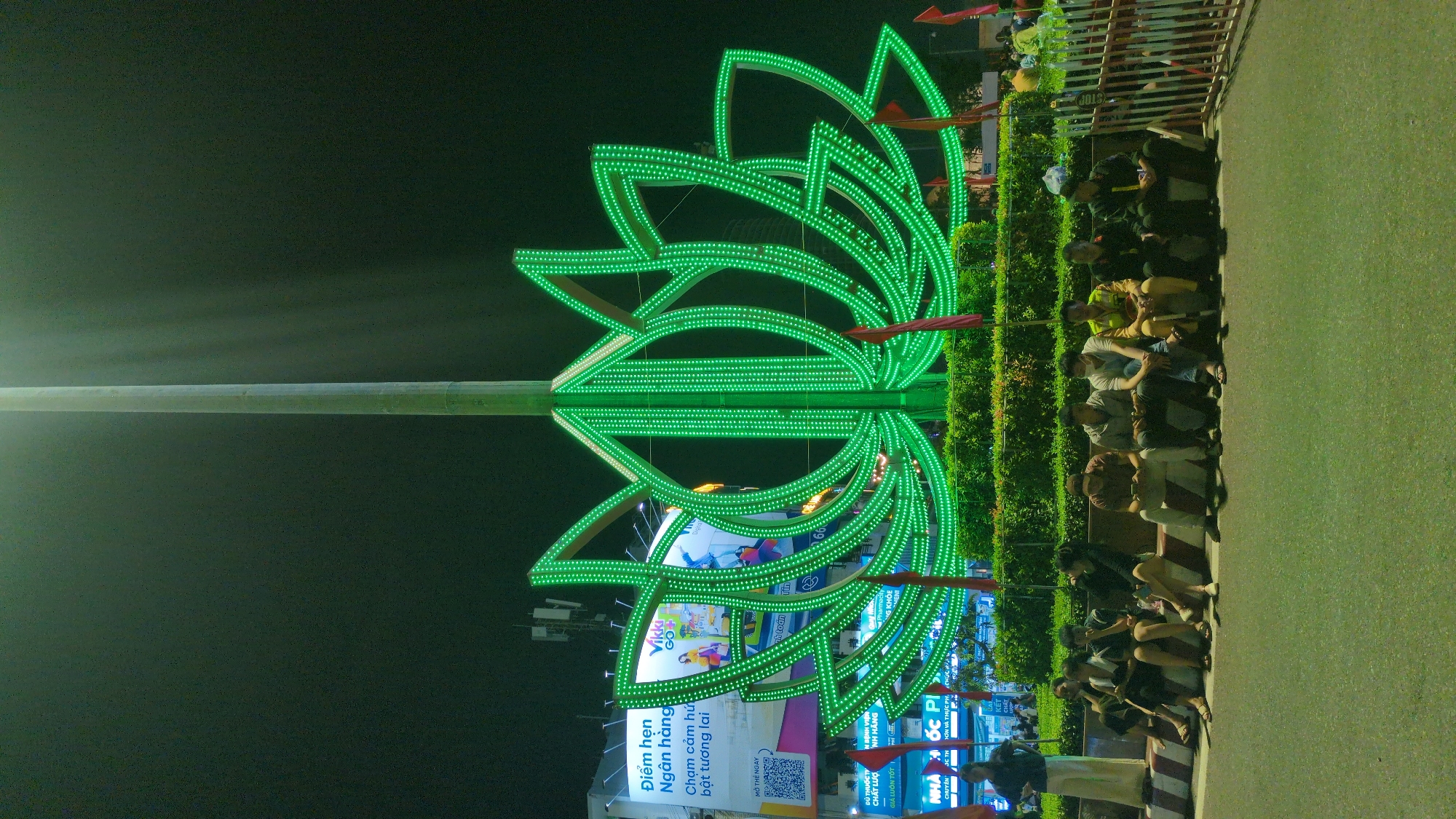 The image captures a nighttime urban scene in South Quy Nhon Ward, Vietnam. A large, green LED light sculpture, stylized as a lotus flower, stands centrally, illuminated against the dark sky. A tall, unlit pole extends vertically from the sculpture's core. The lotus structure is situated on a raised circular median, which is planted with dark green hedges and decorated with several small red flags.
In the background, multi-story buildings display brightly lit billboards with Vietnamese text and imagery, including advertisements for a financial institution (featuring "Ngân hàng" and "Vibbe GO") and a pharmacy ("NHÀ THUỐC PHÚC KHANG").
In the foreground, an asphalt road surface is visible, separated from a pedestrian area by a striped curb (white and reddish-brown). A group of ten individuals is seated along this curb or a low retaining wall. The group consists predominantly of males in casual attire; several appear to be occupied with mobile devices. One individual stands on the far left. On the right side of the seated group, two individuals are distinguishable by uniforms, one in yellow-green and another in dark green. A white and red striped metal barrier is partially visible on the far right.