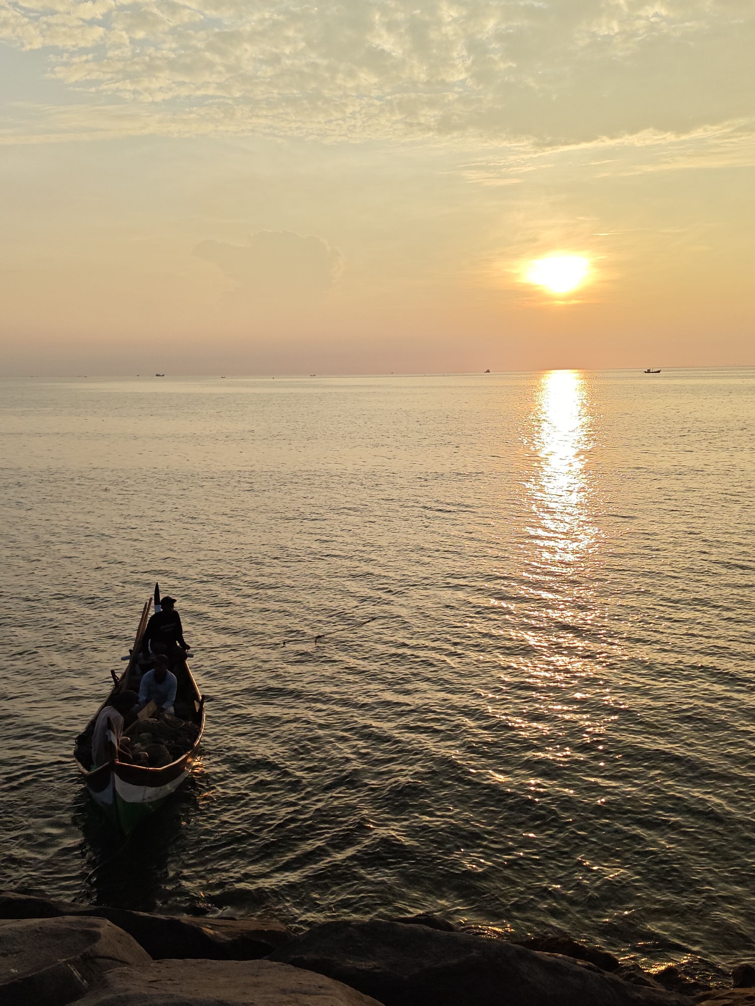The image depicts a sunset over the ocean, likely in the coastal city of Lhokseumawe, Indonesia. In the foreground, a small fishing boat is visible, occupied by several individuals. The boat is positioned near a rocky shoreline. The sun is low on the horizon, casting a bright reflection across the water's surface. Several distant boats are visible along the horizon. The sky is filled with soft, diffused light and clouds, indicating the sun is setting.