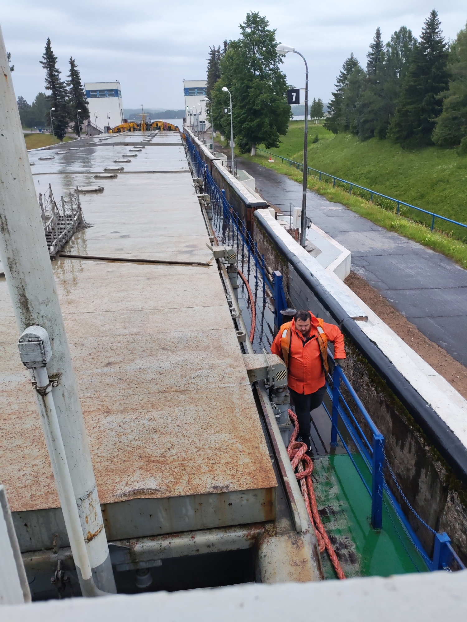 A man wearing an orange life vest and black pants walks along a walkway next to a canal lock. The walkway has a blue railing. A thick, red rope is coiled on the walkway. In the background, there are trees and buildings with white walls and blue trim. The sky is overcast. The location is the Белоусово canal lock, located in Белоусово, Russia.