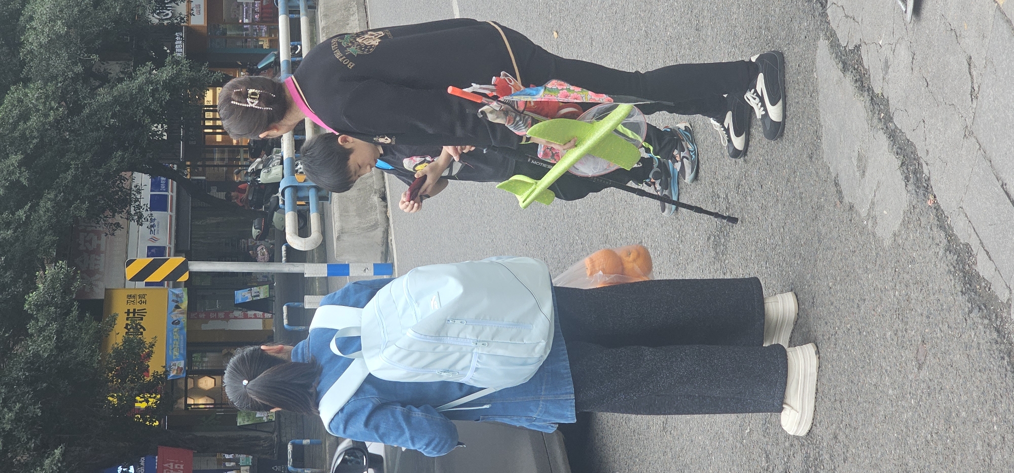 The image captures a street scene in what appears to be Shapingba District, Chongqing, China. A woman with a hair clip and wearing a black sweatshirt is bent over, possibly attending to a child looking at an object. The child appears to be holding something in their hands and is near a stroller with toys. Behind them, another individual with a light blue backpack is visible. The ground is paved, and in the background, one can see buildings with Chinese signage. The presence of trees and street fixtures suggests an urban environment.