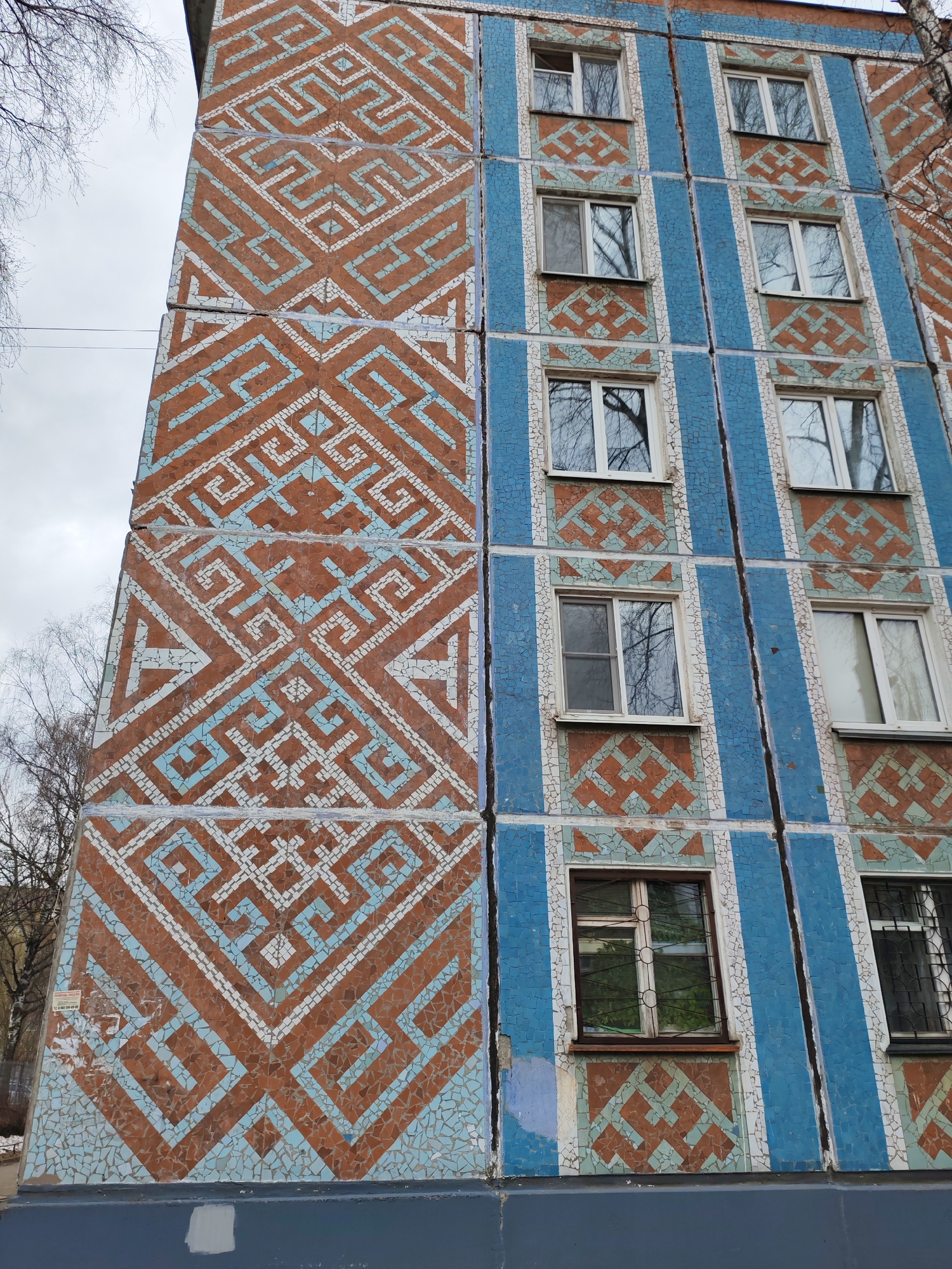 A low-angle view shows a building exterior in Izhevsk, Russia. The building is clad in a mosaic of brown, light blue, and white tiles. The left side of the building features a large geometric pattern. The right side is divided into blue vertical stripes between rows of windows. A brown geometric pattern is visible above and below the windows. The sky is overcast, and bare tree branches are visible at the top of the frame.
