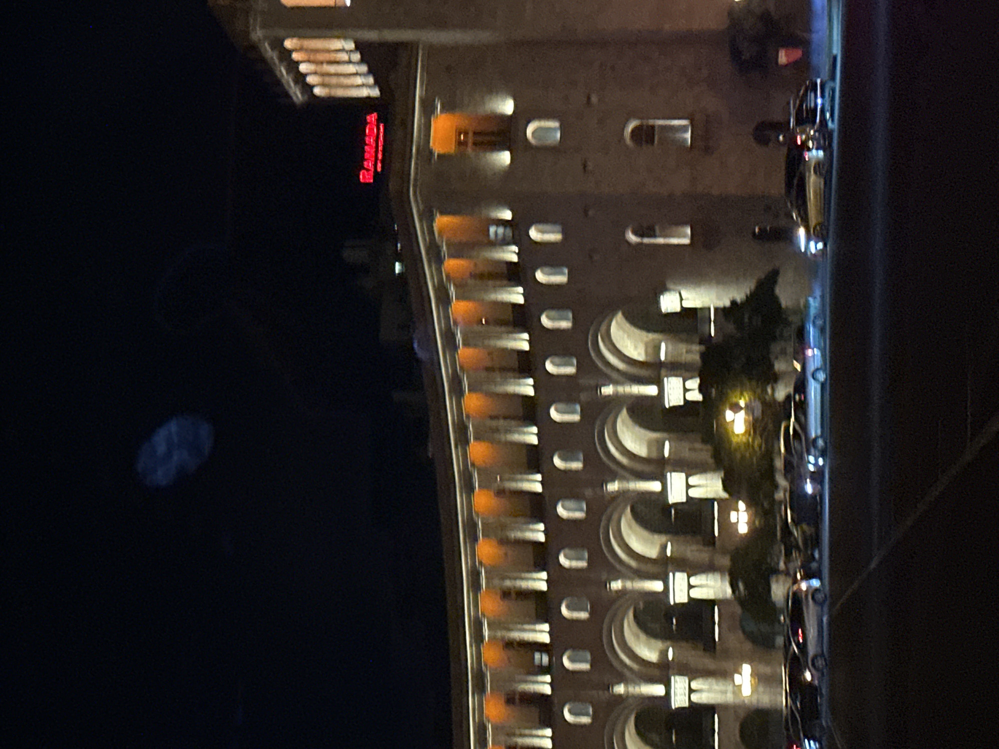 An exterior, eye-level, nighttime, vertical, low-angle shot captures a modern building with illuminated architectural features and a street with parked and moving vehicles. The building, identified by a red "RAMADA" sign at the top, features a curved facade with rows of arched windows and columns, illuminated by orange and white lights. Several vehicles, including cars and SUVs, are visible on the street in front of the building. The sky is dark, with a faint, blurry oval shape visible in the upper left quadrant, possibly an artifact of the camera or an atmospheric phenomenon. The scene is located in Yerevan, Armenia, specifically on Republic Square.