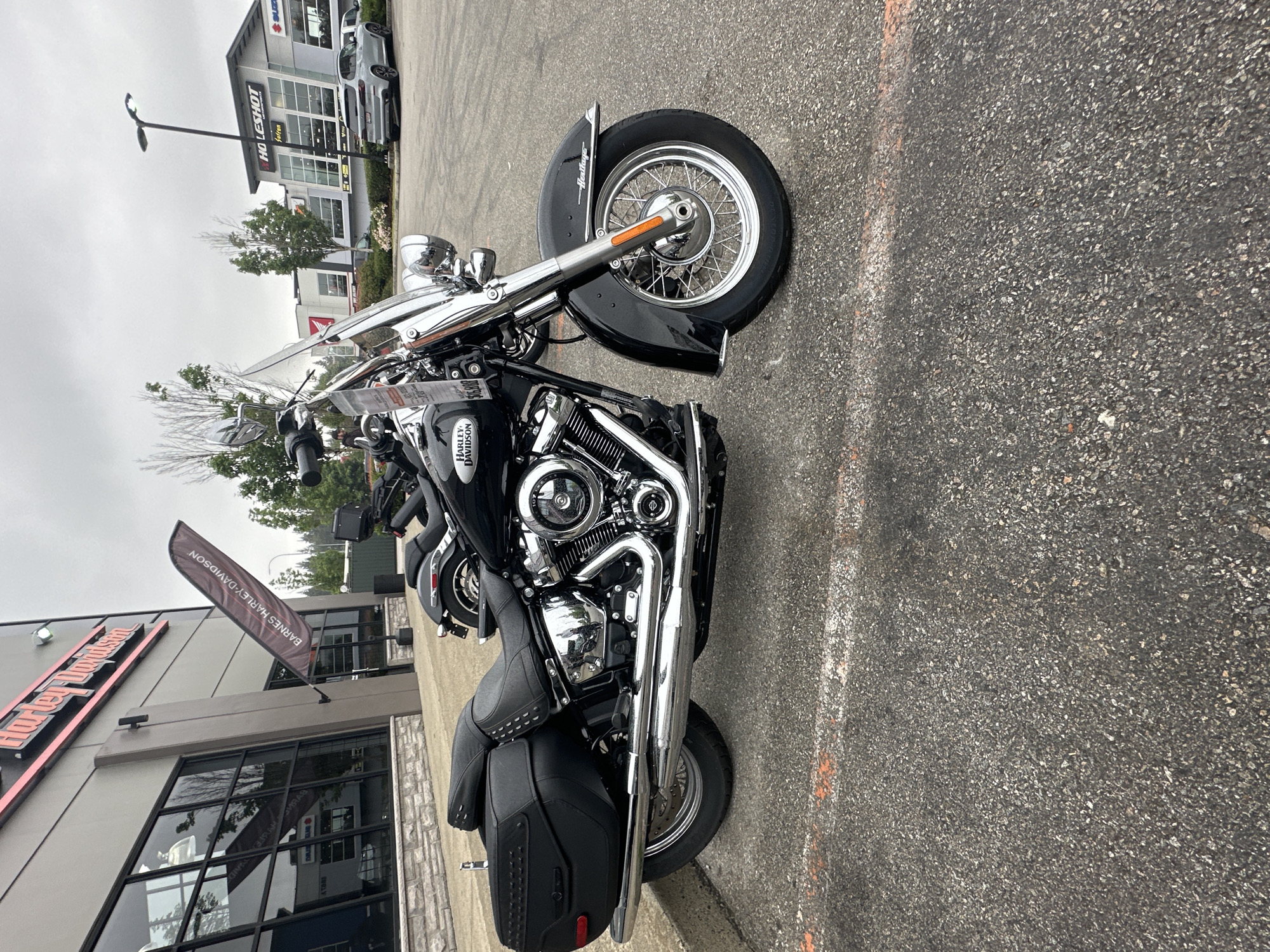 The image shows a black Harley-Davidson Heritage Classic motorcycle parked on asphalt outside a dealership in Township of Langley, Canada. The dealership building, with "Harley-Davidson" branding, is visible in the background. Also in the background is a "Holeshot" dealership and a silver SUV. The sky is overcast. The motorcycle is the primary subject, featuring chrome accents, saddlebags, and a windshield. An orange line is painted on the asphalt in the foreground.