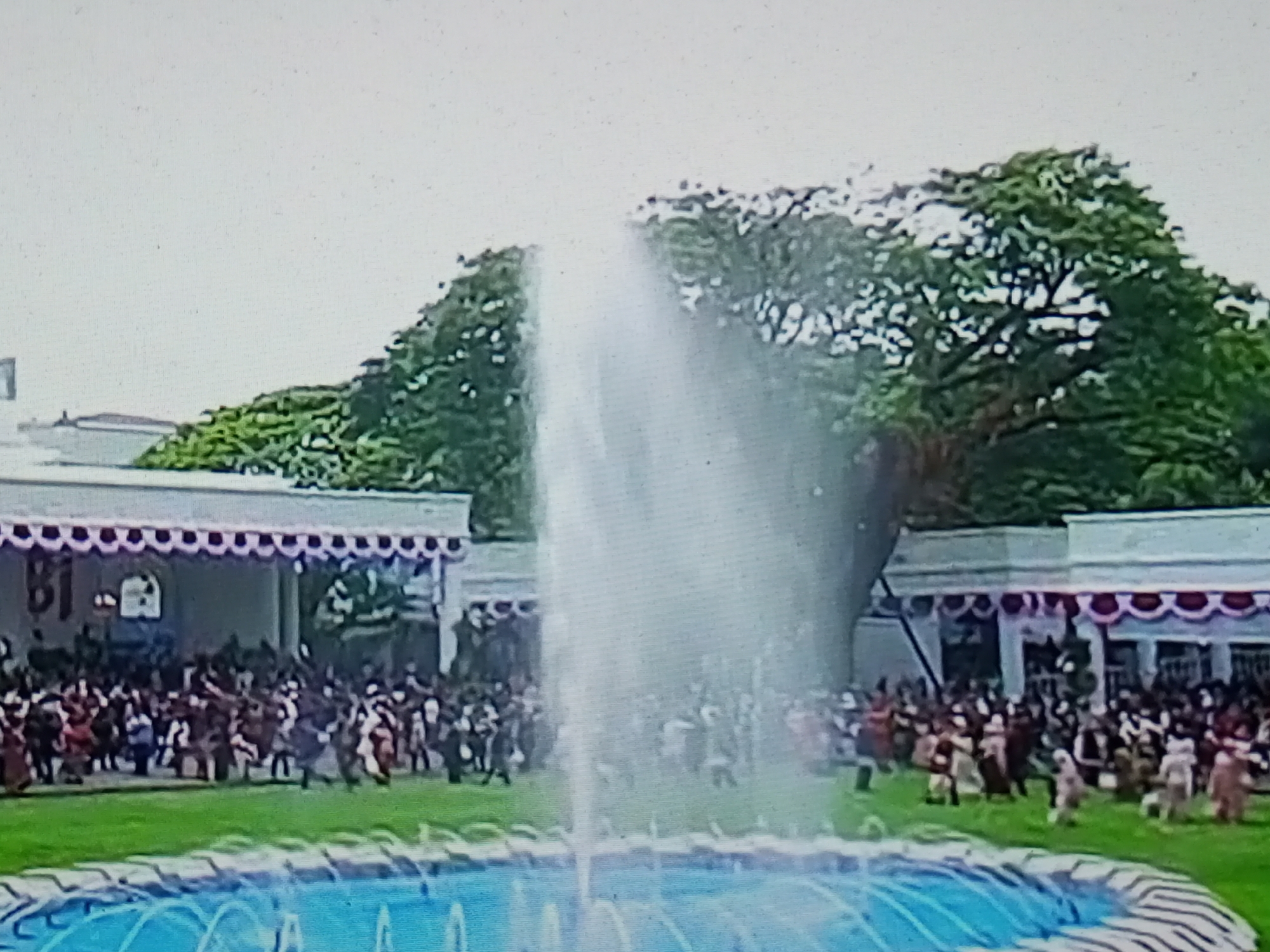 The image shows a crowd of people gathered in an outdoor setting with a large fountain in the foreground. Behind the fountain, there is a long, white building with a decorative red and white trim along the roofline. The building appears to be a pavilion or a part of a larger structure, with columns visible. A lush green lawn extends from the building towards the fountain. Several people are visible on the lawn, some in the distance and others closer to the building, suggesting a public gathering or event. The background features large, mature trees with dense green foliage. The sky is overcast and light-colored. The scene appears to be in Banyudono, Indonesia, likely a public space or a ceremonial ground. The fountain is actively spraying water, creating a dynamic element in the center of the frame. The people in the crowd are dressed in various attire, some in what appears to be traditional clothing, suggesting a cultural or festive occasion.