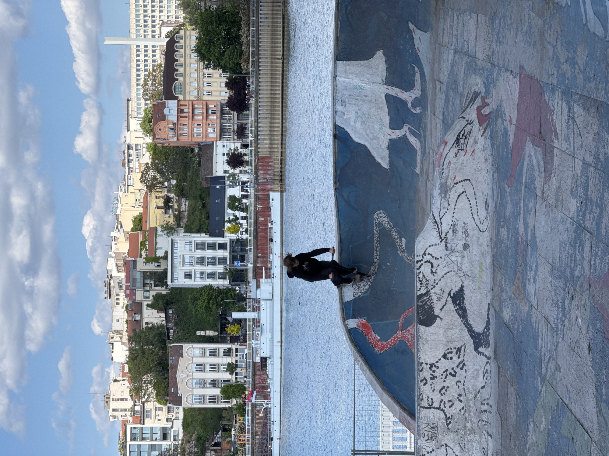 The image captures a vibrant daytime scene in Belgrade, Serbia. In the foreground, a person is seen standing on a skateboard, balancing on the edge of a curved concrete structure that appears to be part of a skatepark or a riverside embankment. This structure is richly decorated with colorful, abstract murals featuring stylized white, black, and red shapes that resemble figures or creatures. The individual is dressed in dark clothing, including a hoodie and pants.
Behind the skateboarder, a wide body of water, likely the Sava River, stretches across the middle ground. A white vessel or structure on the far bank distinctly shows the word "SAVA". On the opposite bank, a dense cityscape unfolds under a bright blue sky dotted with scattered white clouds. The skyline features numerous multi-story buildings of various architectural styles and colors, including older residential blocks and more modern structures, interspersed with green trees. A tall, modern white building is prominent in the background. The scene depicts a lively urban environment where art, recreation, and city life converge by the river.