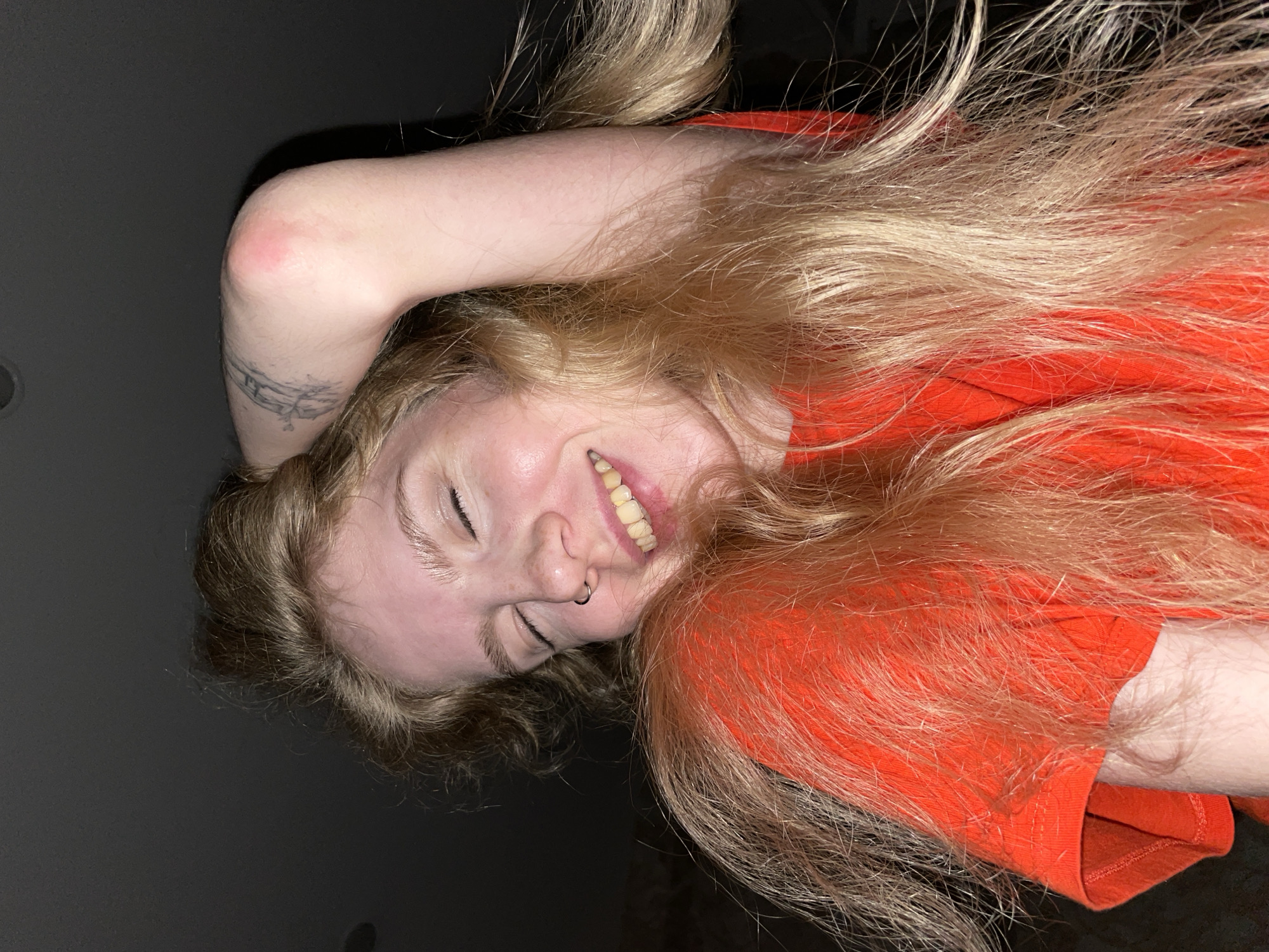 A young woman with blonde hair and closed eyes smiles with her arm raised behind her head, revealing a tattoo on her arm and a red mark on her elbow. She is wearing an orange t-shirt. The background is dark, suggesting an indoor environment. The scene is lit from above. There is no information in the image to suggest a location in Izhevsk, Russia.