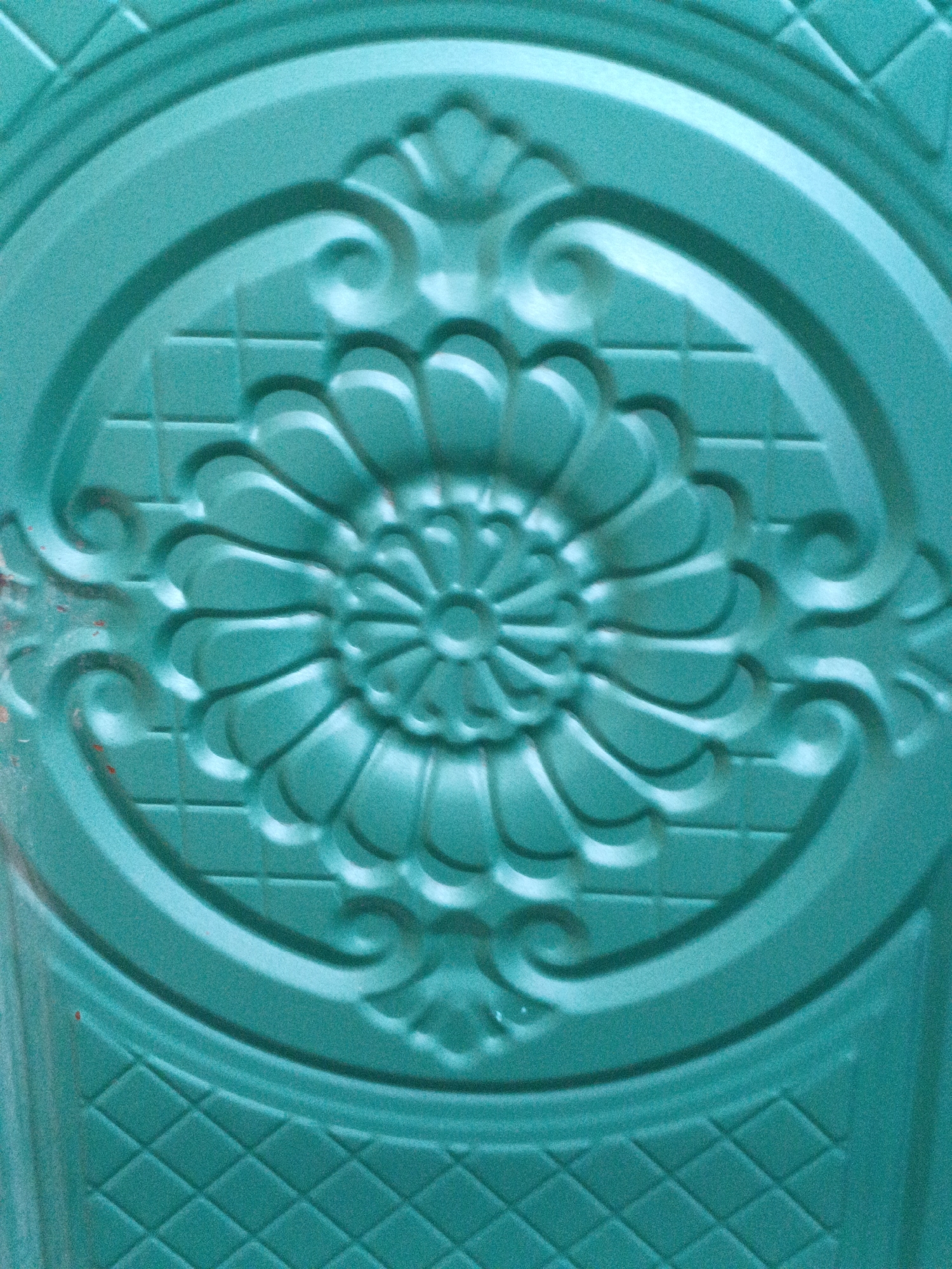 The image shows a close-up of a decorative, embossed metal surface, likely a door or gate, with a turquoise patina. The central design features a floral motif within an oval frame, surrounded by scrolling filigree. The background exhibits a subtle diamond-shaped pattern. There are no people or discernible actions depicted. The location is not specified within the image or its metadata.