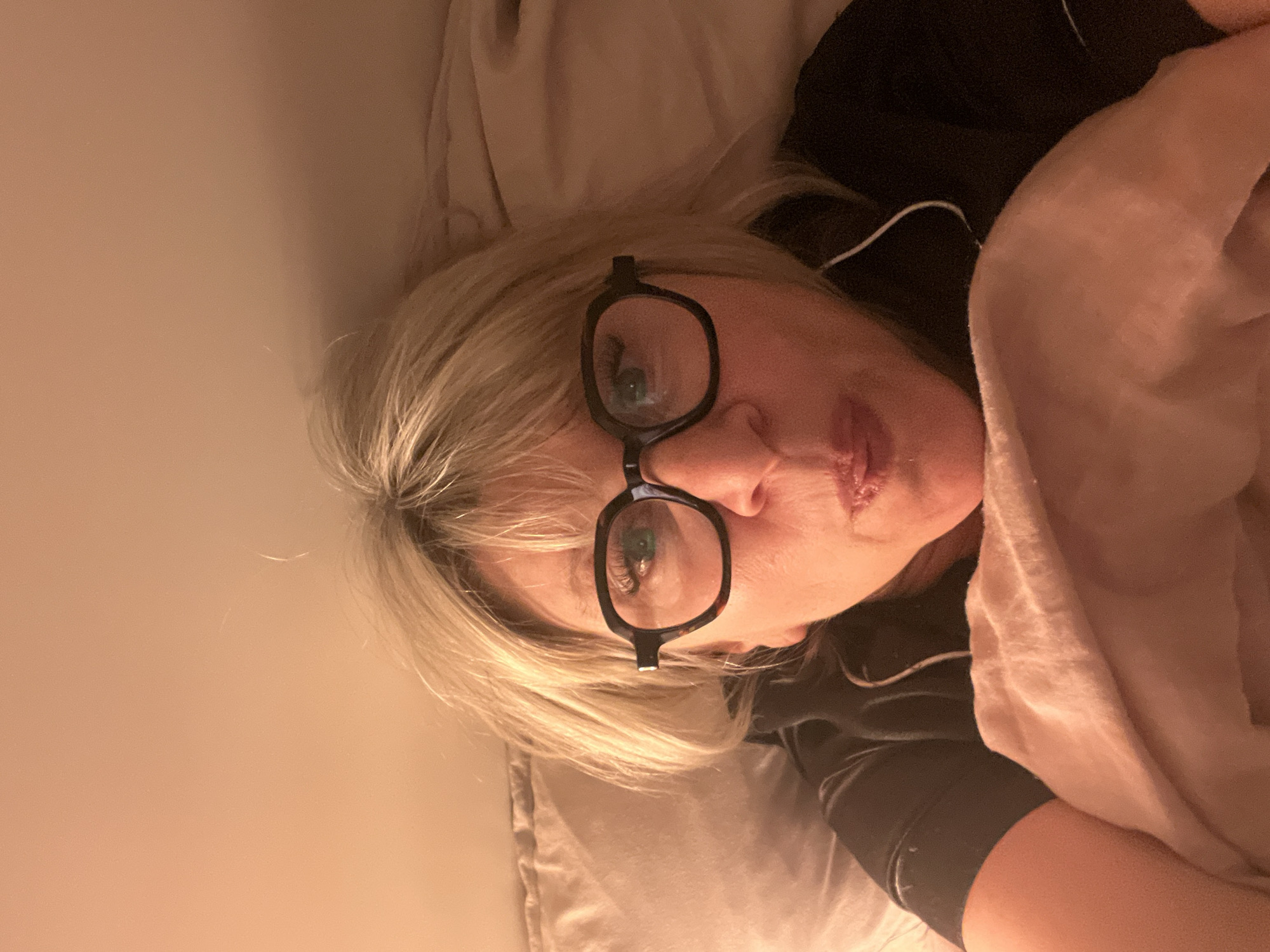 A middle-aged woman is lying in bed. She has blonde hair, fair skin and is wearing eyeglasses. She is wearing black pajamas with white trim. She is covered with a light pink blanket and is lying on a beige pillow. There is a plain wall behind her. The location is unknown and the image does not necessarily depict the city of Chambœuf, France.