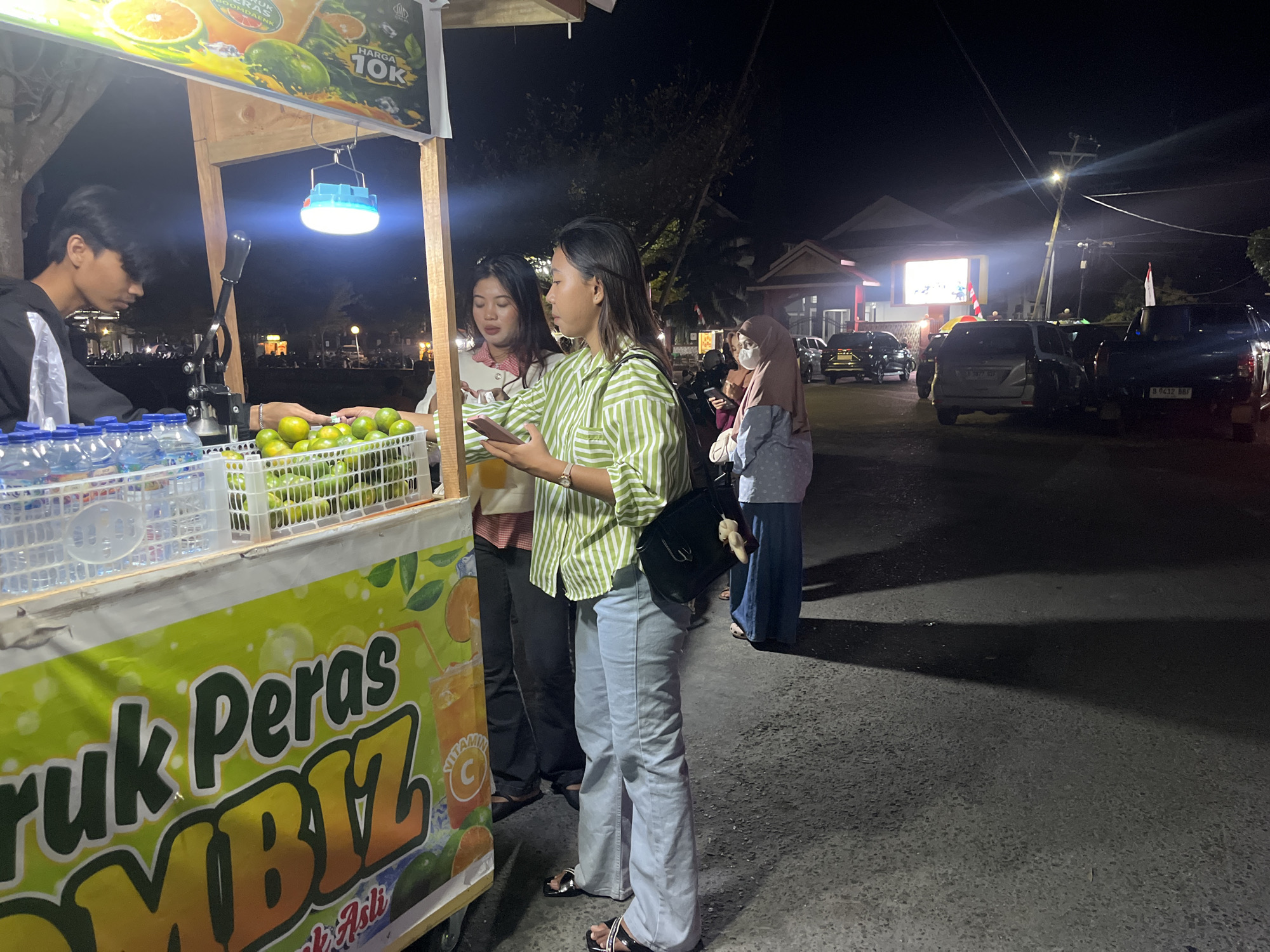 In Luwuk, Indonesia, a street vendor at night is serving a customer at a juice stand. The stand is illuminated by a blue LED light. The vendor, a young male in a black hoodie, is behind the counter, which displays bottles of water and a basket of oranges. A young female customer, wearing a green and white striped shirt and light blue jeans, is paying for her order, holding a smartphone in her hand. Another female customer, wearing a white shirt with a red and white checkered collar, stands next to her. In the background, several cars and buildings are visible. A large screen displays an image. The overall scene depicts a bustling night market.