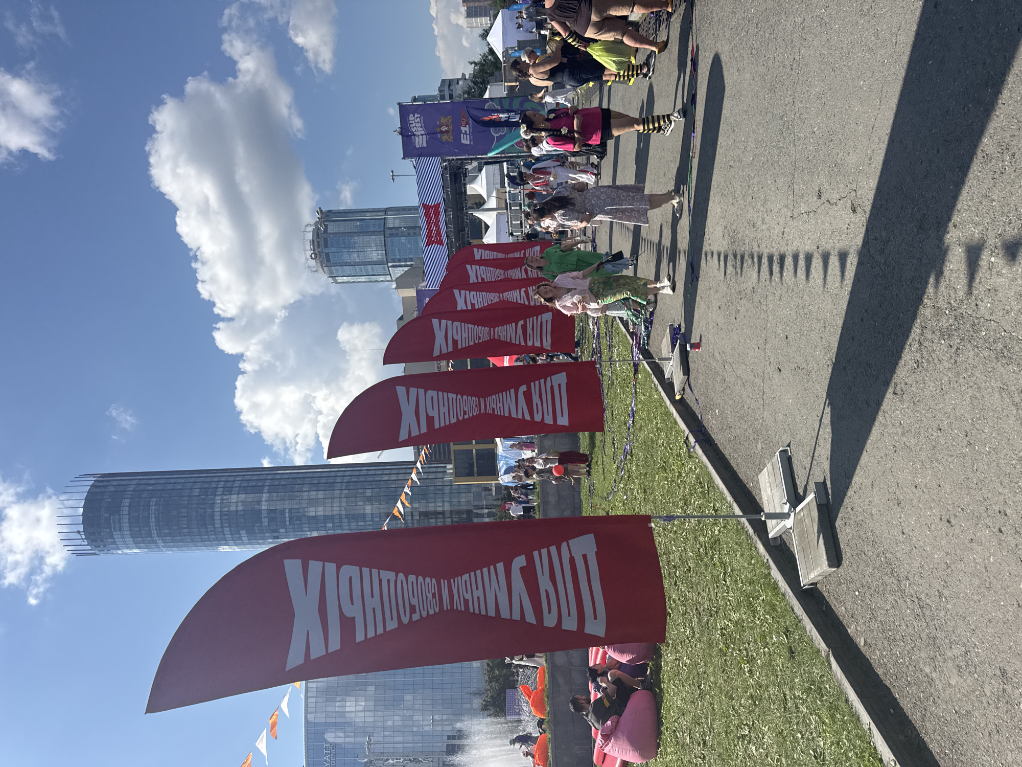 The image depicts an outdoor festival or event taking place on a sunny day in Yekaterinburg, Russia, near the Hyatt Regency hotel. In the foreground, several large red flags with white text are prominently displayed, standing on poles. The text on the flags appears to be in Russian and is oriented vertically. One of the flags reads "ДЛЯ УМНЫХ И СВОБОДНЫХ" (For the Smart and Free). Behind the flags, people are milling around on a paved area and a grassy section.
On the grassy area, several large, inflatable beanbag chairs are visible, with some individuals seated on them. A fountain is also present in the background on the left.
In the middle ground, a procession of people are walking along a paved path. They are dressed in casual attire. Further back, there are white tents and arches, suggesting various event areas or stages. The iconic Yekaterinburg skyscraper, Iset Tower, is visible in the background, along with other modern buildings, indicating an urban setting. The sky is blue with scattered white clouds. The overall atmosphere appears festive and lively.