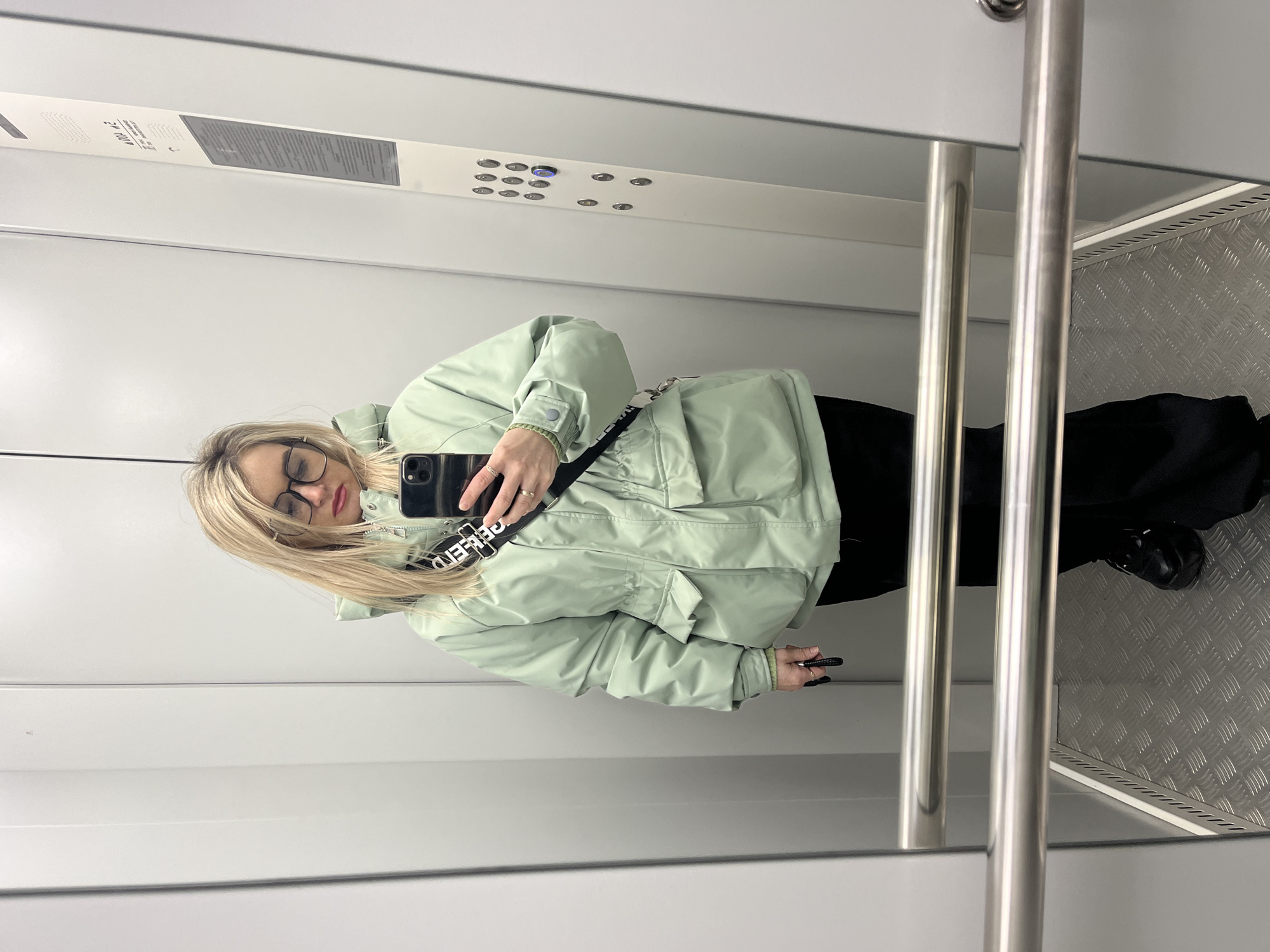 A person stands in an elevator cabin, presumably in Chaikovsky, Russia, although this is not directly discernible from the image. The person is wearing a light green puffer jacket, black trousers, and black shoes. They have blonde hair and glasses. They are holding a smartphone, seemingly taking a selfie in the elevator mirror. The elevator's interior consists of grey walls, a control panel with buttons, stainless steel handrails, and a textured metal floor.