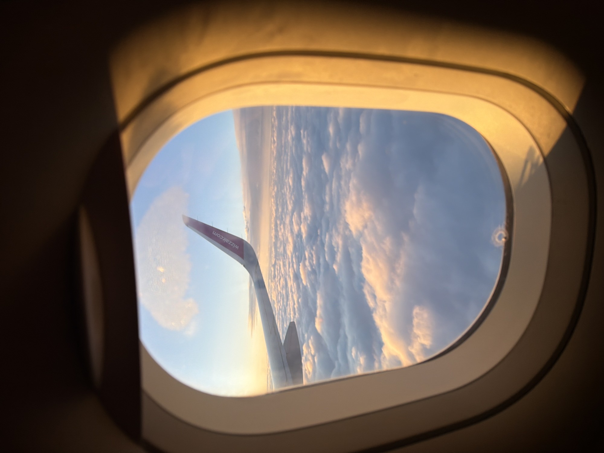 An airplane wing, identifiable by the purple logo "wizzair.com" on its tip, extends into the frame from the left, soaring above a vast expanse of fluffy white clouds illuminated by the warm, golden light of a sunset or sunrise. The view is framed by the interior of an airplane window, with its characteristic rounded oval shape and layered, cream-colored trim. The surrounding interior of the cabin is dark, with the light from outside casting an orange glow on the window frame. The scene is devoid of people and any urban or Czerwony Kościół specific environmental details.