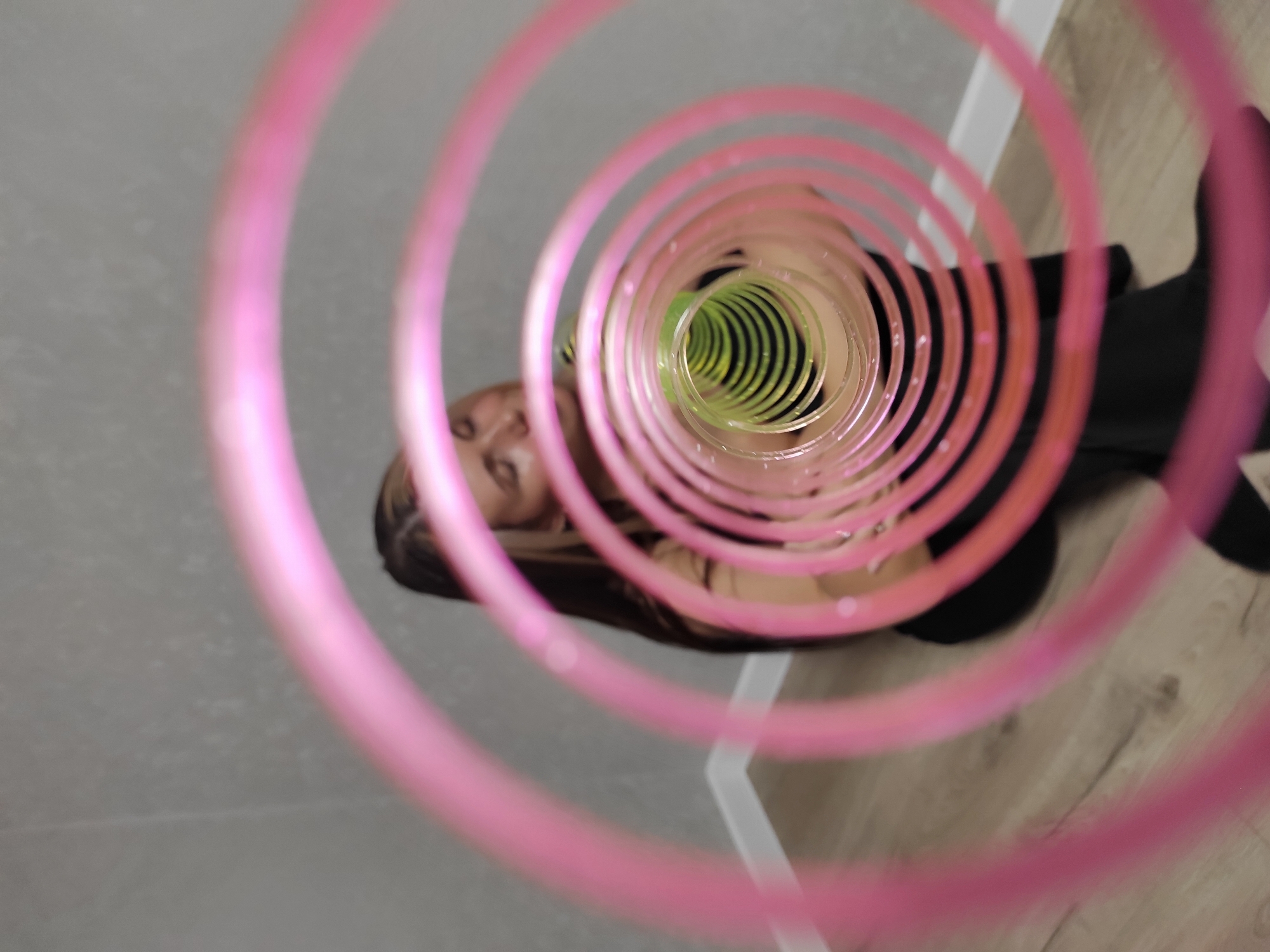 A high-angle shot shows a young woman's face as seen through the opening of a partially collapsed, multi-colored, plastic slinky toy. The slinky is pink at the top, transitioning to green in the interior coils. The woman has long brown hair and is wearing a black top. The background consists of a gray tile floor and a white baseboard. A partially visible wood floor is also in view on the right side of the image. The image was taken in the city of Chaikovsky, Russia.