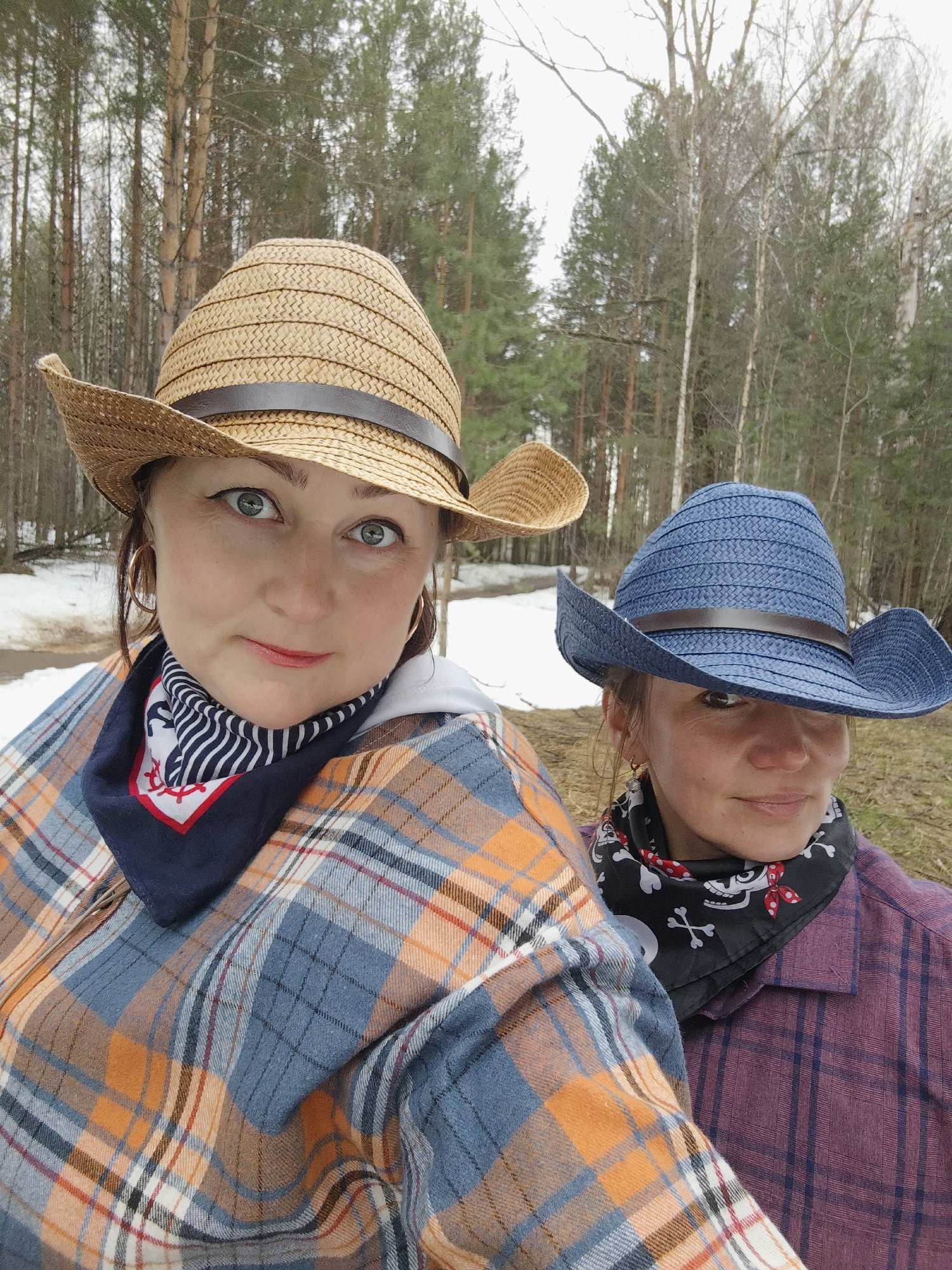 Two women are posing for a selfie outdoors, possibly in a wooded area in the city of Chaikovsky, Russia. The woman in the foreground is wearing a light-brown straw cowboy hat, an orange and blue plaid shirt, and a navy-blue bandana with white stripes and a red logo. The other woman, behind, is wearing a blue straw cowboy hat, a purple plaid shirt, and a black bandana with skull and crossbones. The environment includes bare trees and patches of snow on the ground.