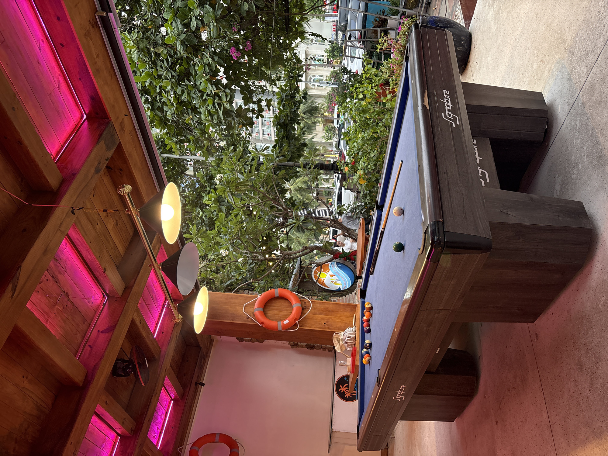 A well-lit, outdoor bar area in Đà Nẵng, Vietnam, features a billiard table with balls arranged at one end, a cue stick, and a single ball near the center of the blue felt. The table, branded "Signature", is positioned under a partial wooden structure with pink LED strip lights and a three-headed adjustable spotlight. A life preserver and a tropical-themed sign are mounted on the wall behind the table. In the background, through the foliage, there is a view of a street with vehicles and buildings.