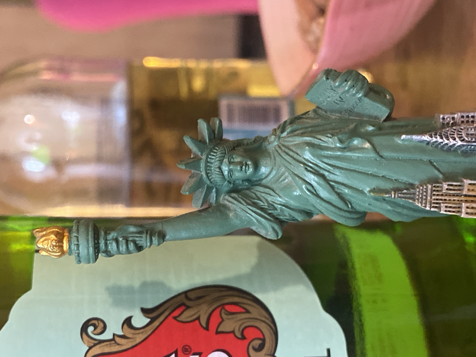 A close-up photograph depicts a souvenir statue of the Statue of Liberty, with its signature green patina and golden torch, resting on what appears to be a green bottle with a red and brown label. The location is not specified, therefore I can't say if it is in the city of Votkinsk, Russia. The background includes blurred images of other items, including what may be a bottle and a pink object.