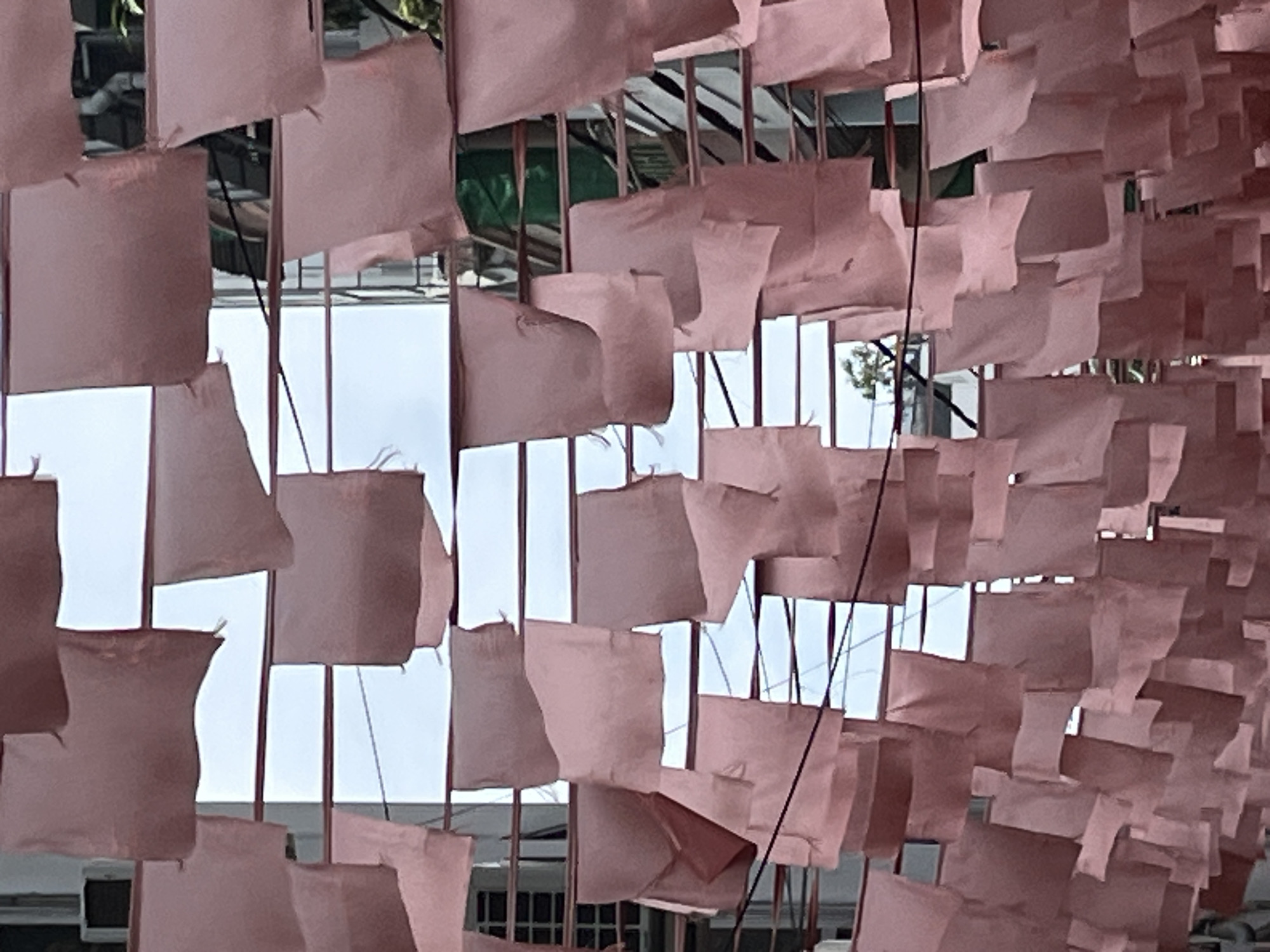 The image shows a dense arrangement of pink fabric squares hanging from horizontal lines, creating a canopy effect. The fabric pieces vary in size and are hung in multiple rows, overlapping each other. They are unevenly spaced and some appear to be creased or folded, suggesting they are lightweight and affected by air currents. Behind the hanging fabric, a building structure with horizontal and vertical elements is visible, appearing to be made of metal or a similar material. The sky, visible through the gaps in the fabric and structure, is overcast or pale. The scene suggests an outdoor installation or decoration. The location of Nha Trang Ward, Vietnam, suggests a possible cultural or festive context for this display.