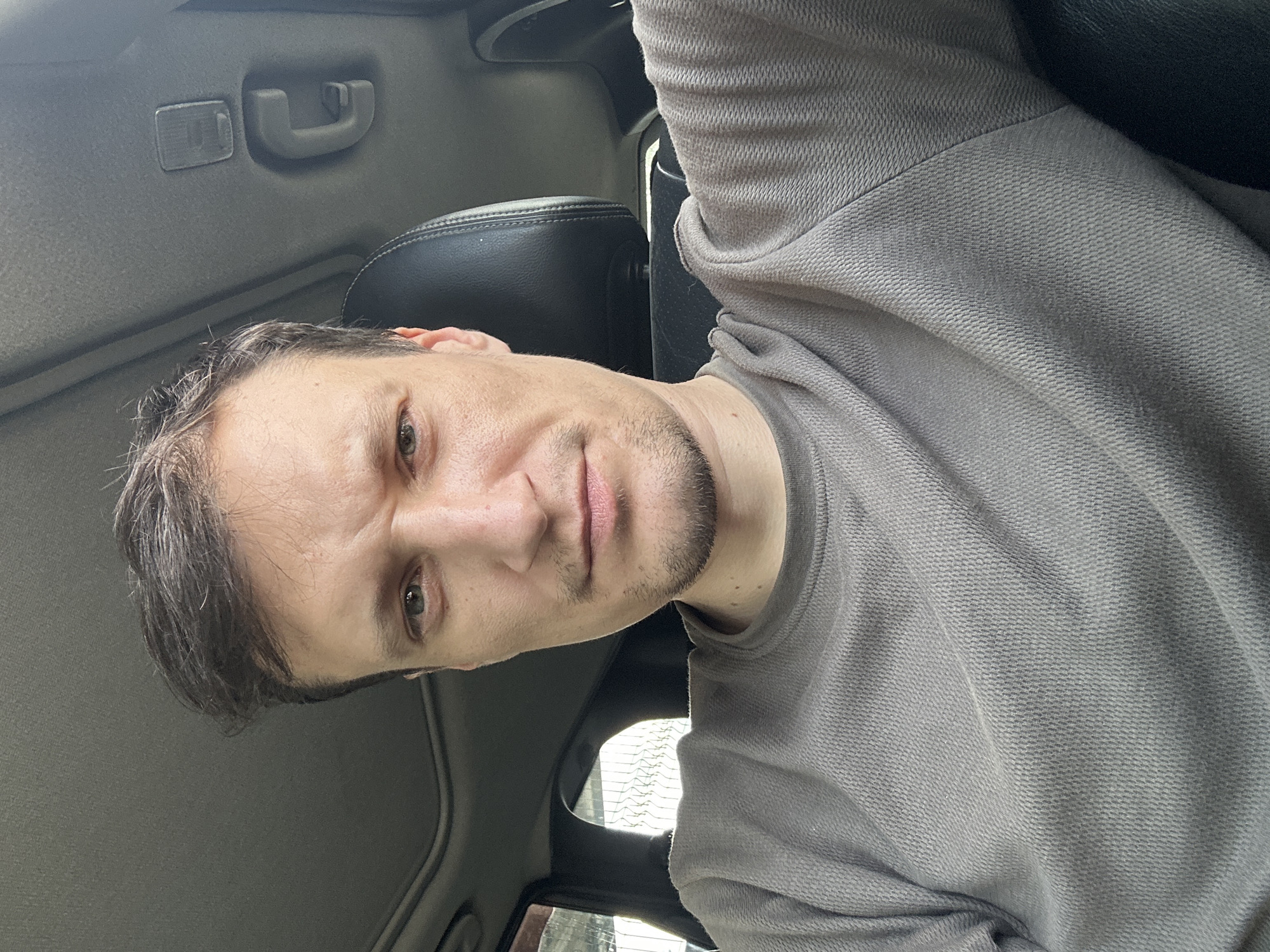A man with short, dark hair and a beard is sitting in the driver's seat of a car, looking at the camera. He is wearing a grey long-sleeved shirt. The car's sunroof is open, and the interior of the car, including a headrest and part of the dashboard, is visible. The scene appears to be outdoors in Shatava, Ukraine.