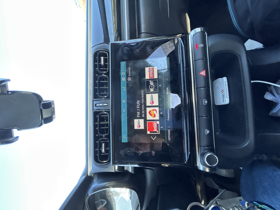 The scene depicts the interior of a vehicle, located in Saint-Jean-Bonnefonds, France. The perspective is from the front passenger area, looking towards the dashboard and windshield. A large, rectangular infotainment screen dominates the central dashboard, displaying "FM / FUN" with "96.30 MHz FUN RADIO," alongside logos for radio stations "inter," "SCOOP," "MOUV'," and "SKYROCK." The screen indicates a current time of 11:10 and an external temperature of -2°C. A reflection of a human face, consistent with a young male with short dark hair, is visible on the left side of the infotainment screen. Above the screen are four horizontal air vents. Below the screen, a row of tactile buttons includes controls for defrost and rear defroster, with an illuminated red hazard light triangle indicating active hazard lights, and an illuminated red lock icon indicating the doors are locked. In an open storage compartment beneath these controls, a white 'VINCI' telepass device, a white cable, and crumpled white paper are present. A keyring with multiple keys and a blue accessory is visible on the lower left. Part of a person's leg, clad in blue jeans, is visible on the lower left, while dark fabric is visible on the lower right. Through the windshield, a clear, bright sky is visible, with a black phone holder mounted at the top of the glass. A distant, partial blue structure is discernible on the far right horizon.