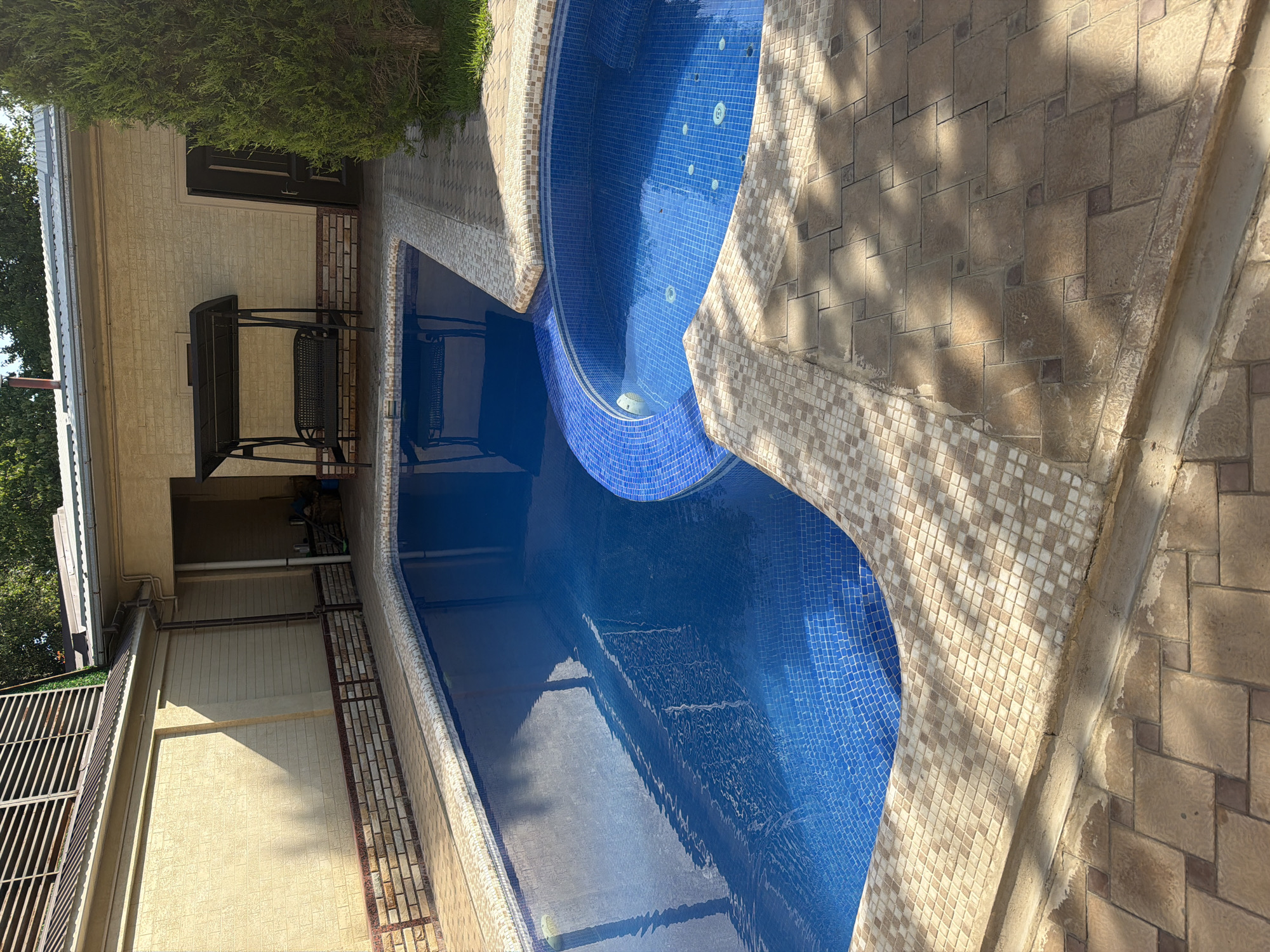 The image depicts an outdoor swimming pool and hot tub area, likely in Koshkurgon, Uzbekistan. The pool is a vibrant blue, tiled with small mosaic squares, and has a curved edge. Adjacent to the main pool is a circular hot tub, also tiled in blue mosaic. The surrounding area is paved with tan and brown interlocking stones, with a decorative border of smaller beige and tan tiles along the pool's edge. A section of the pool area is shaded by the shadow of what appears to be a covered seating area or pergola with a bench swing. To the right, a tall, green coniferous tree stands near a light-colored building with a dark door. The building has beige textured walls and a metal roof. The overall scene suggests a private residential or recreational space.
