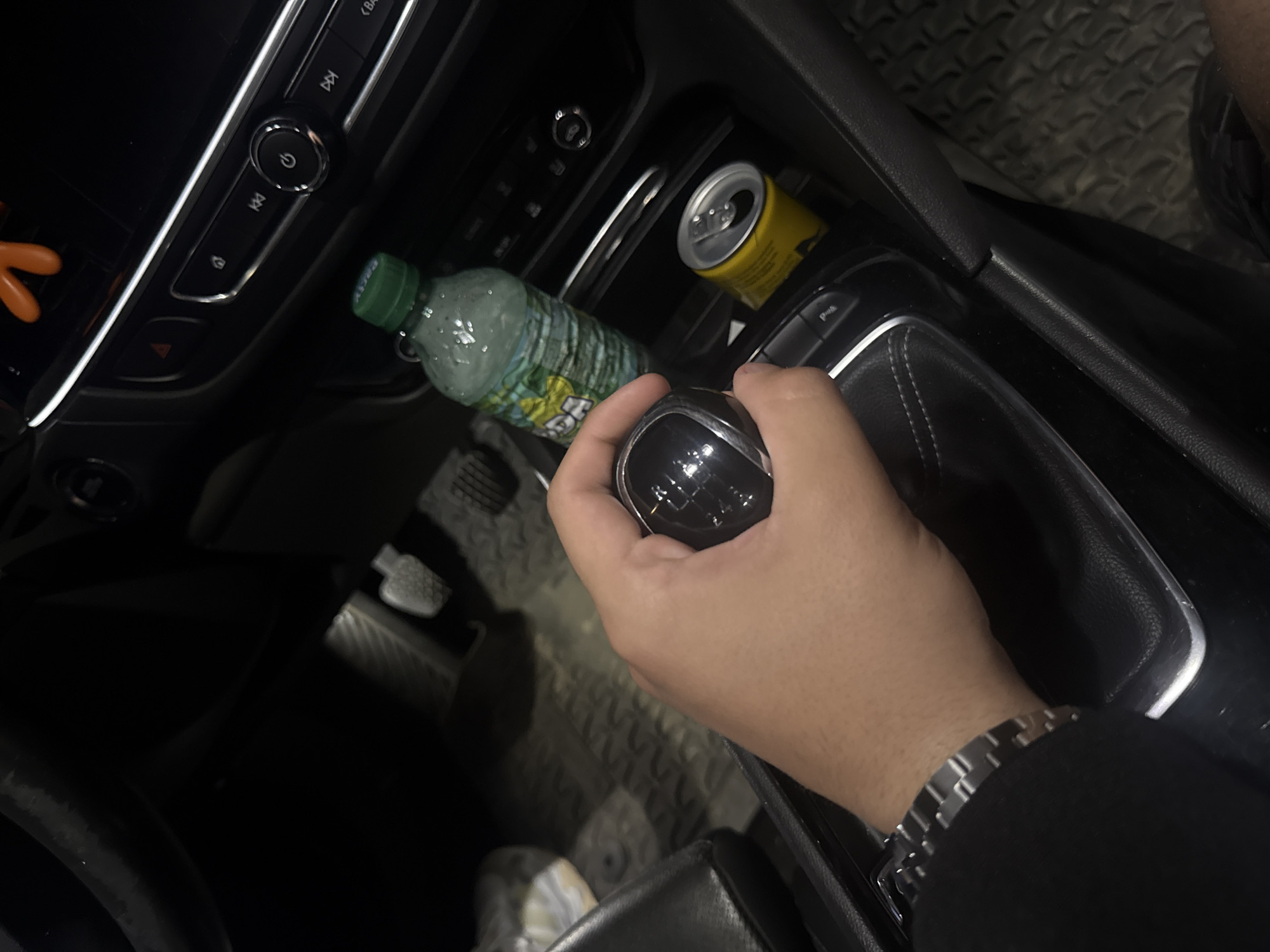 The image depicts the interior of a vehicle located in Varna, Bulgaria, captured from an elevated perspective looking downwards into the footwell and center console area. A light-skinned human hand, wearing a dark long-sleeved garment and a silver-toned metal link bracelet on the left wrist, is positioned around a black manual gear stick with a chrome base. The gear stick's knob displays a 6-speed shift pattern (R-1-2-3-4-5-6).
To the right of the gear stick, within a cup holder, an open yellow aluminum beverage can is present next to a clear plastic bottle with a green cap. The bottle, featuring condensation, has a partially visible green label with the word "SODA" and contains a light-colored liquid.
The center console and dashboard area exhibit various dark-colored controls with chrome accents. Visible buttons include media navigation controls, a circular power button, and a red triangular hazard light button. Two foot pedals, consistent with a manual transmission, are discernible on the dark, patterned rubber floor mat in the lower-left background. A section of a black seat is also visible on the left side of the frame. The overall environment is dimly lit.