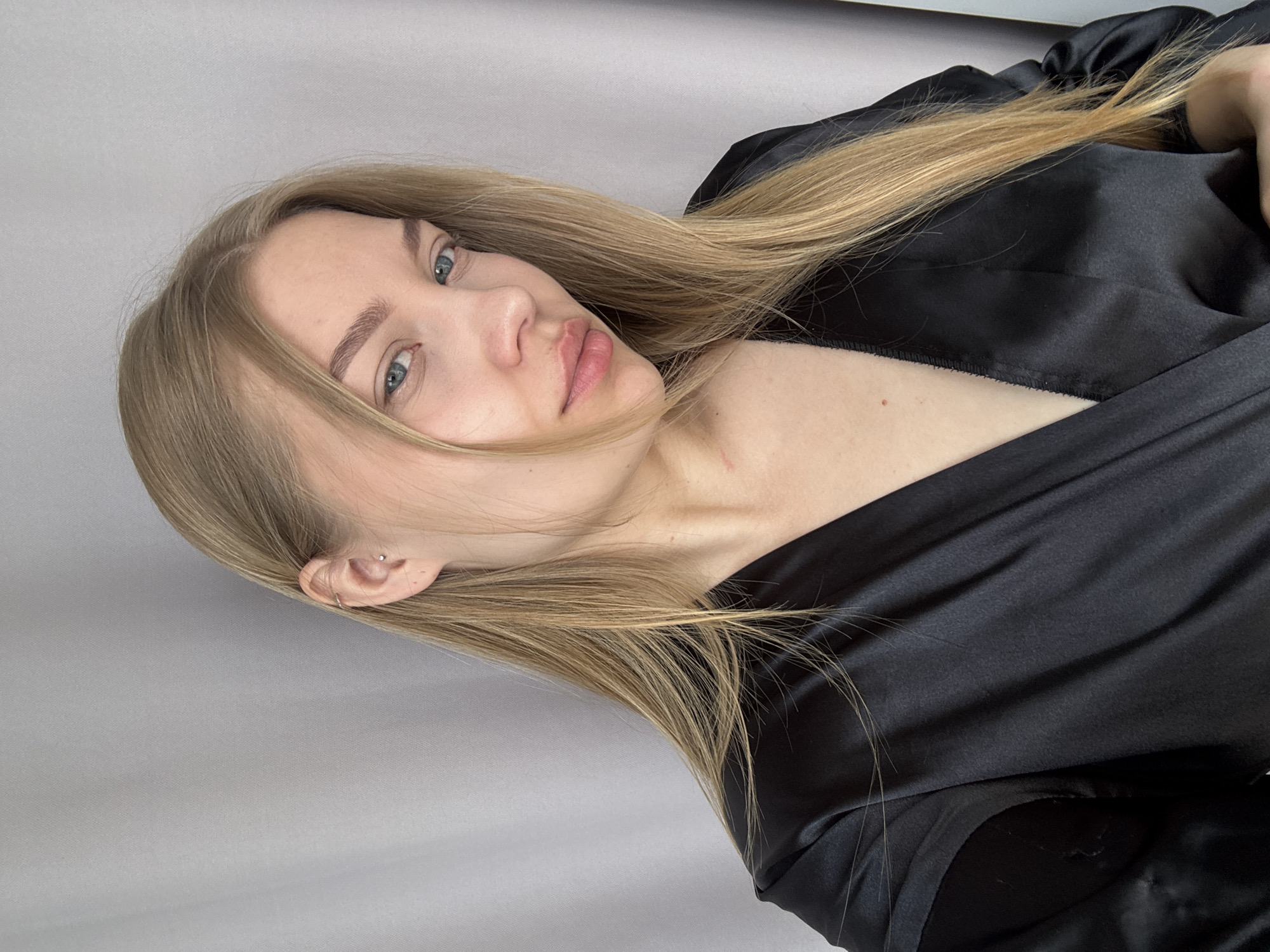 A close-up portrait shows a young woman with long, straight, blonde hair and light blue eyes. She is positioned as if reclining or lying down, looking directly at the camera with a calm and neutral expression, her lips slightly parted. Her blonde hair cascades around her, with some strands resting on her shoulder and the black garment she is wearing.
She is dressed in a black, satin or silk-like garment, possibly a robe or a blouse, which drapes loosely around her, revealing her bare shoulder and upper chest. A small, simple silver stud earring is visible in her left ear. A tiny mole is present on her upper chest.
The background is a plain, light grey surface, suggesting an indoor setting, likely a wall or a studio backdrop. The lighting is soft, diffused, and even, illuminating her face and the textures of her hair and clothing without harsh shadows. There are no other discernible objects or visible text in the image. The overall atmosphere is serene and intimate. While the context indicates the image was taken in Chaikovsky, Russia, the scene itself offers no specific geographical cues, nor does it provide information about the time of day or weather.