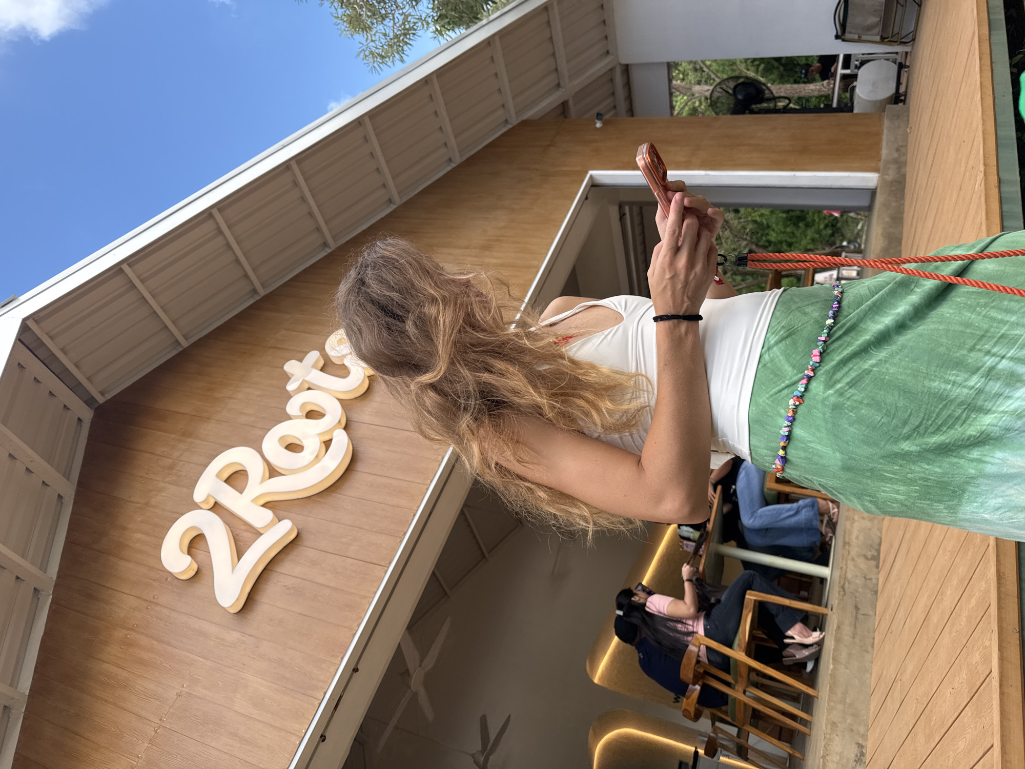 A woman with long, light brown hair is seen from the side, holding a copper-colored smartphone in both hands, possibly taking a photo. She is wearing a white tank top and a green patterned sarong. A bracelet is visible on her left wrist. To her left is a building with the sign "2 Roots" in large, stylized lettering. Below the woman and slightly to the right, two people are seated at tables, suggesting an indoor or semi-outdoor dining area. The image is taken during the day, with a clear blue sky visible in the background. The location is Kuta Selatan, Indonesia.