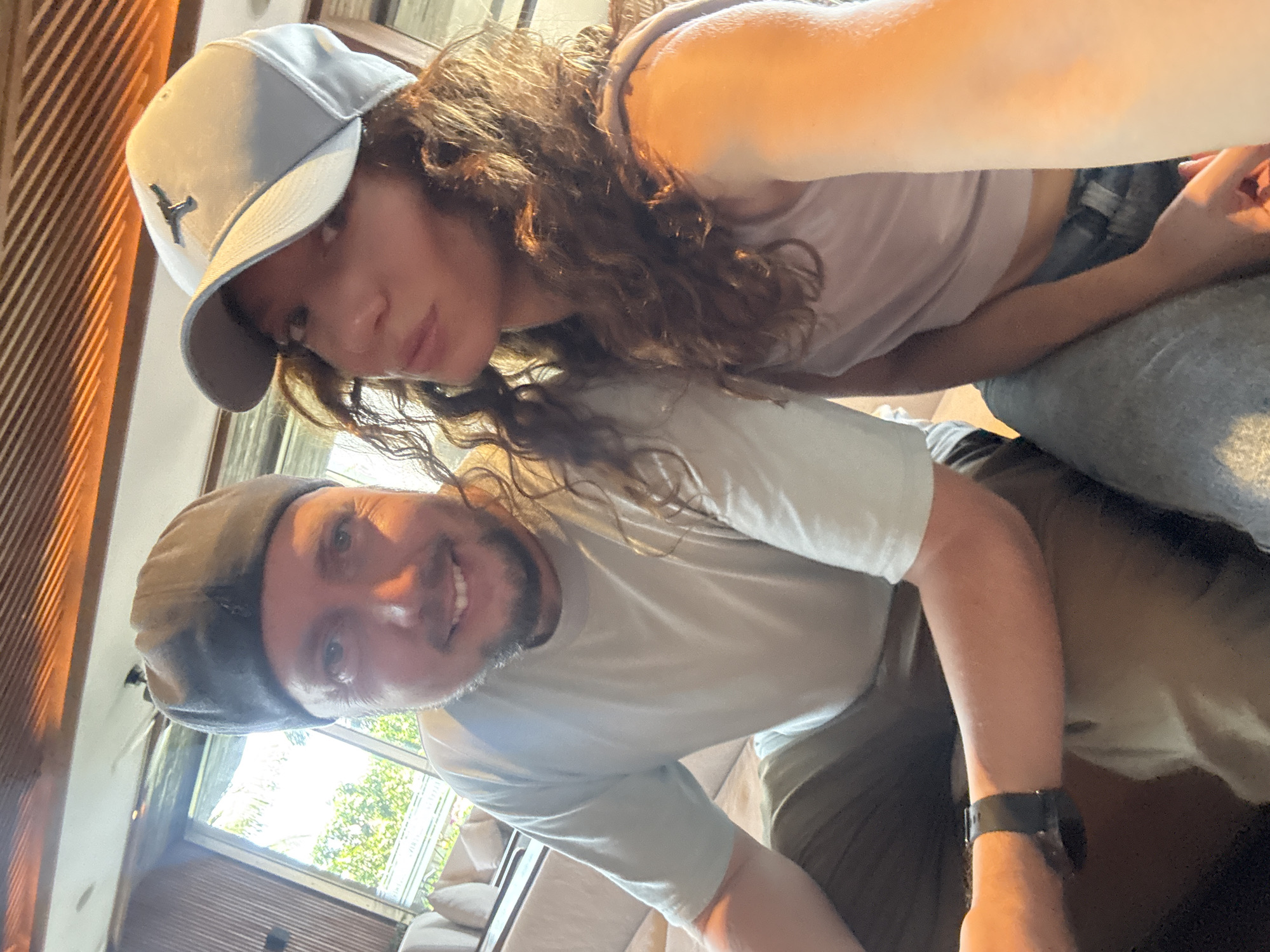 Two people are posed close together, likely taking a selfie. The woman wears a light-colored baseball cap with a logo and a light-colored top. The man wears a dark cap, light-colored shirt and a watch on his left wrist. A window with trees visible outside is in the background. The scene appears to be indoors in a living space with furniture and wood-paneled walls. The location in Đà Nẵng, Vietnam cannot be determined from this image.