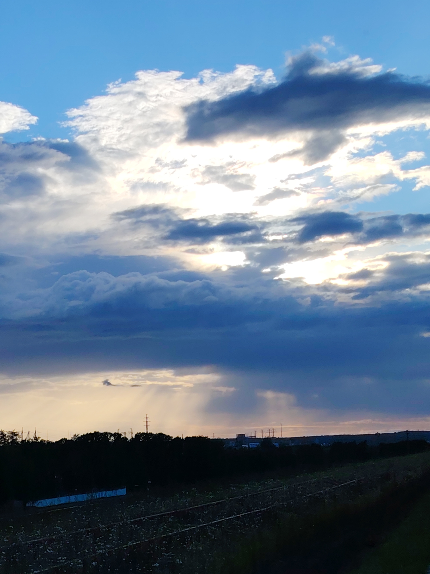 This image captures a dramatic sky over a rural landscape in Chaikovsky, Russia. The sky is filled with a dynamic array of clouds, ranging from bright white, sunlit formations to dark, imposing storm clouds. Rays of sunlight pierce through the clouds, creating a luminous and somewhat ethereal effect. Below the sky, a dark, silhouetted line of trees forms the horizon, behind which various industrial structures and power lines are visible, suggesting a semi-rural or industrial fringe setting. The foreground consists of a dark, grassy area with what appear to be railway tracks or some form of linear infrastructure, possibly an agricultural field with rows. The overall mood is one of transition, perhaps hinting at an approaching storm or the fading light of day. The time of day appears to be late afternoon or early evening, given the warm tones in the sky and the dramatic lighting. The weather seems to be changing, with both bright sunshine and dark clouds present. There is no visible text in the image.