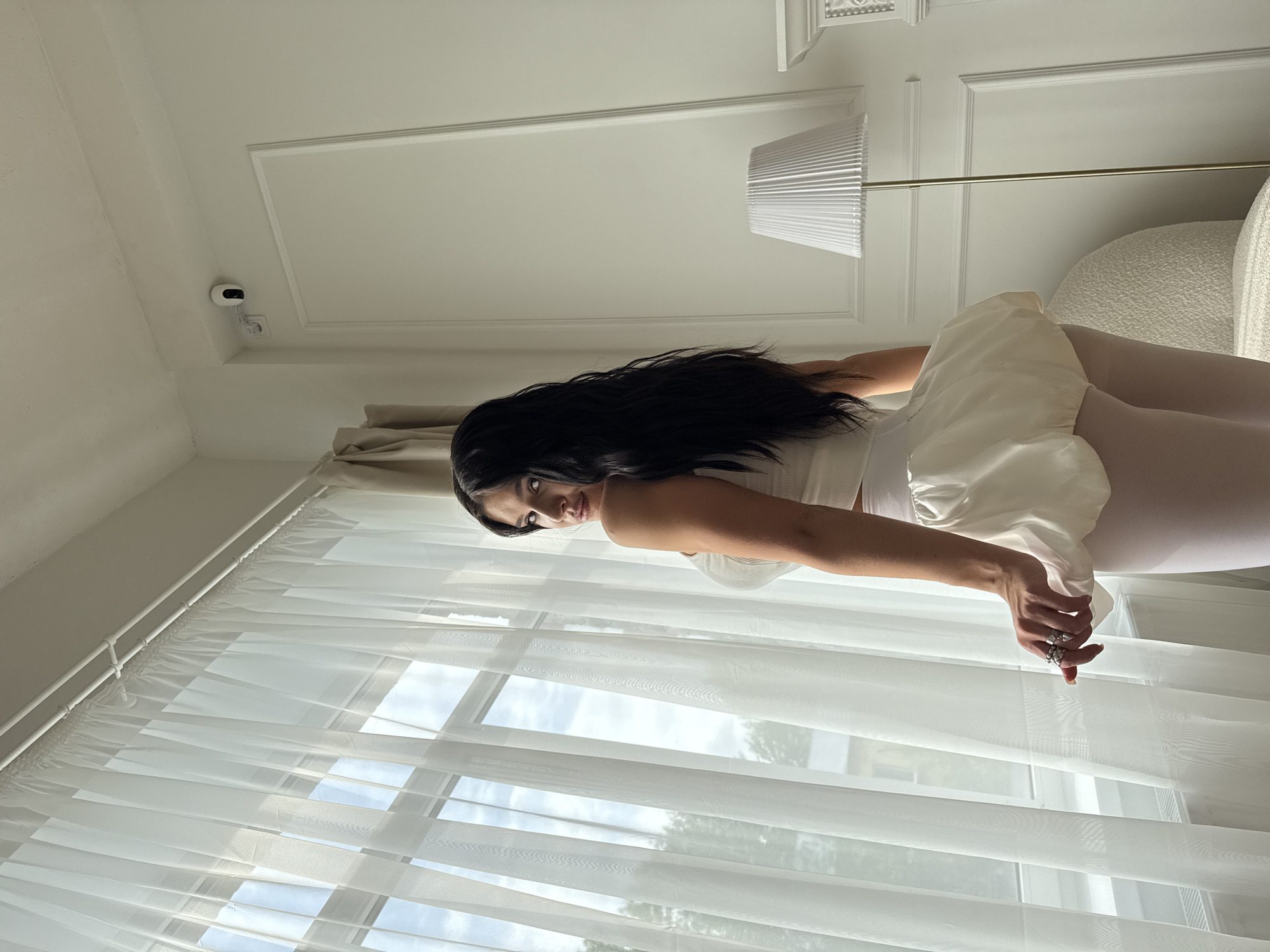 A woman with long, dark hair is posing indoors. She is wearing a light-colored outfit consisting of a tank top, short shorts, and leggings. She is positioned against a backdrop of a window covered with white, sheer curtains. Outside, there is a view of a daytime sky and trees. The room is decorated with white walls and has a lamp in the background. A security camera is visible on the ceiling. The specific location in Kazan, Russia, cannot be determined from this image alone.