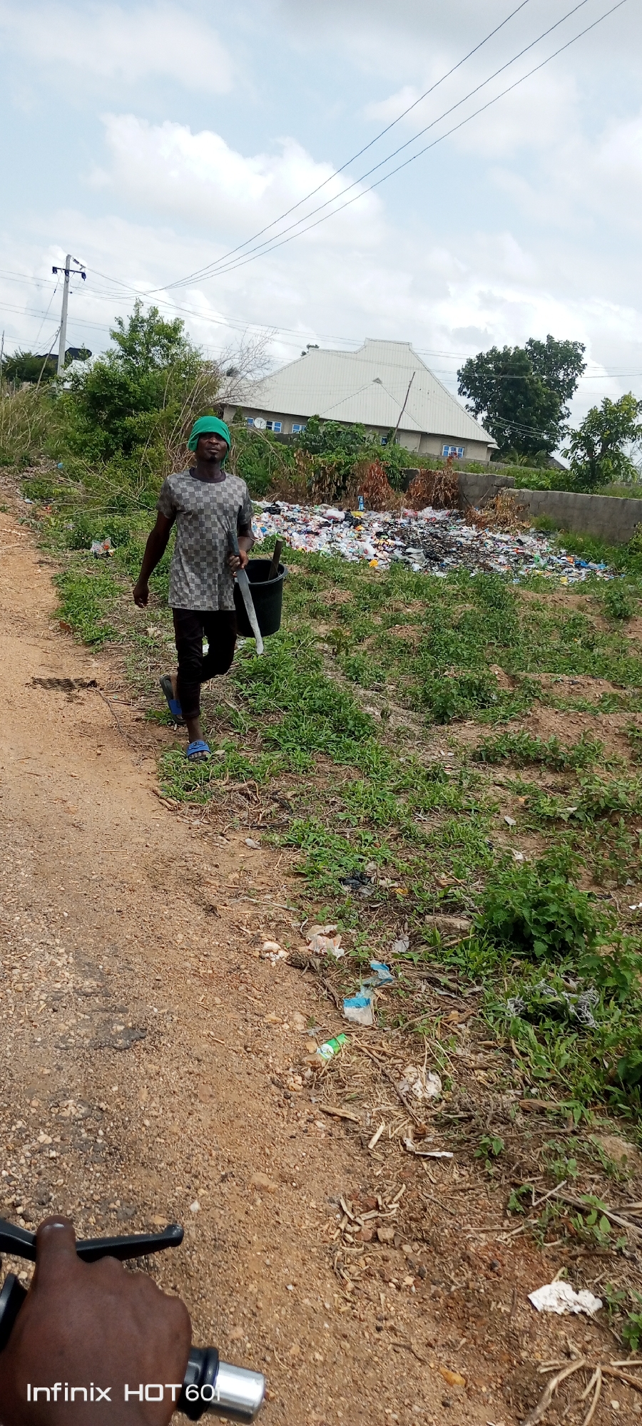 A man in Zango-ndaji, Nigeria walks along a dirt road carrying a bucket and machete. He is wearing a green head covering, checkered shirt, dark pants, and blue sandals. To his right is a large pile of garbage next to a building and vegetation. The foreground shows the handlebars of a bicycle.