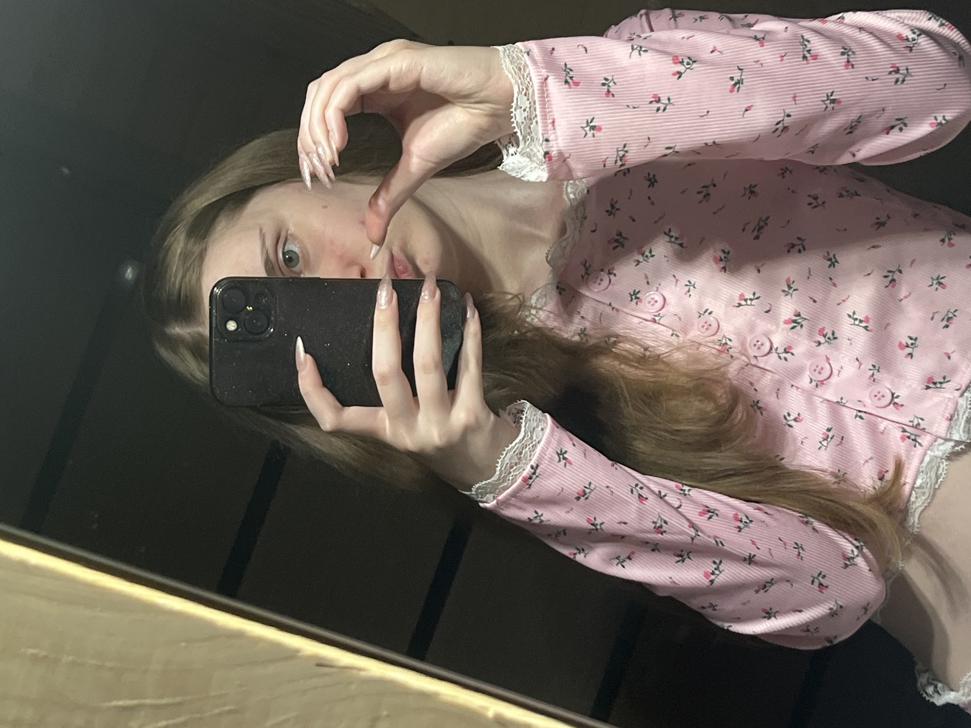 A young woman with long blonde hair takes a mirror selfie. She is wearing a pink, long-sleeved shirt with a floral pattern and white lace trim. Her left hand holds a black smartphone, obscuring part of her face. Her right hand is near her face, with long, light-colored fingernails. The background appears to be an indoor setting, possibly a room with a wooden closet or furniture visible to the left, and a dark, out-of-focus background behind her. There is no indication of the city of Ashkadar, Russia, in the image.