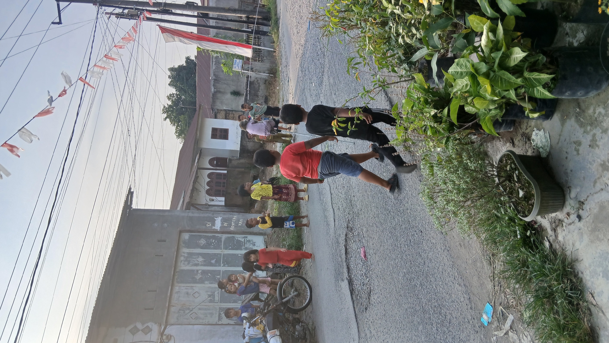 In Tanjung Gusta, Indonesia, a group of children are observed in a residential street scene. Two boys, one in a red t-shirt and blue shorts and the other in a black t-shirt and black pants, walk away from the viewer towards the right. Several other children are gathered on the left side of the street, some standing and observing, others appearing to be interacting. One child is being carried by an adult. A motorcycle is parked near a building on the left. Red and white decorative banners are strung across utility lines overhead, suggesting a possible local celebration or holiday. The environment appears to be a moderately developed urban or semi-urban area, with simple buildings and overhead electrical wires. Potted plants are visible in the foreground on the right. The ground is paved with asphalt, showing some signs of wear and tear. The overall atmosphere is casual and typical of a neighborhood gathering on a clear day.