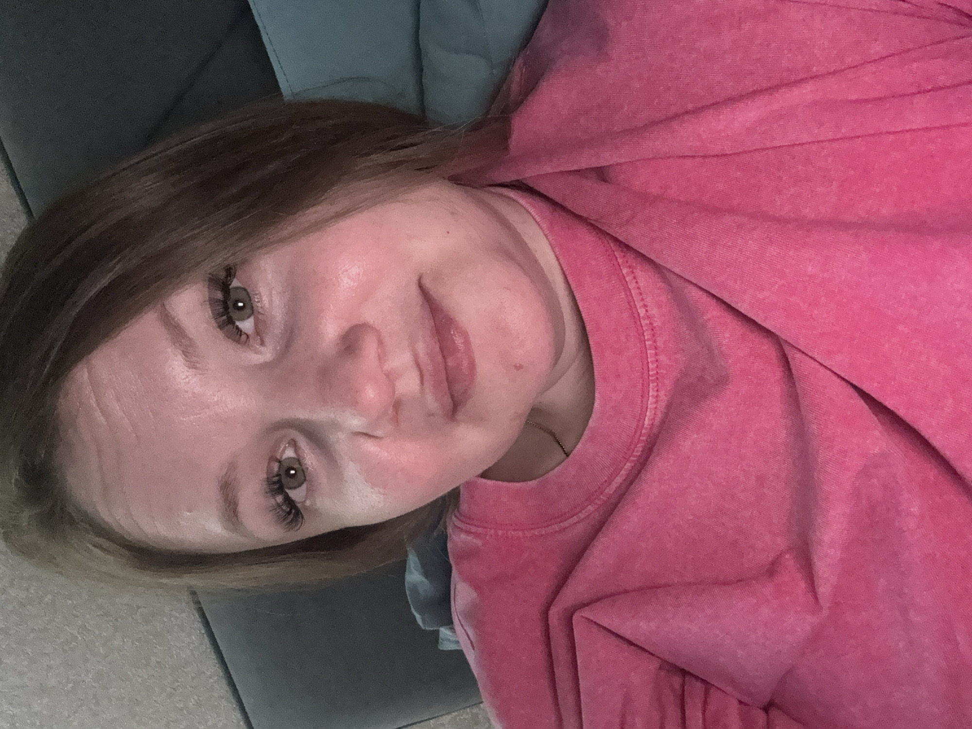 A woman with light skin and hazel eyes, enhanced with false eyelashes, is lying on a gray-blue upholstered surface, possibly a sofa or bed. She is wearing a pink, textured sweatshirt. The background is indistinct, with a gray floor visible. The photograph appears to have been taken indoors. The image does not depict a location in the city of Chaikovsky, Russia.