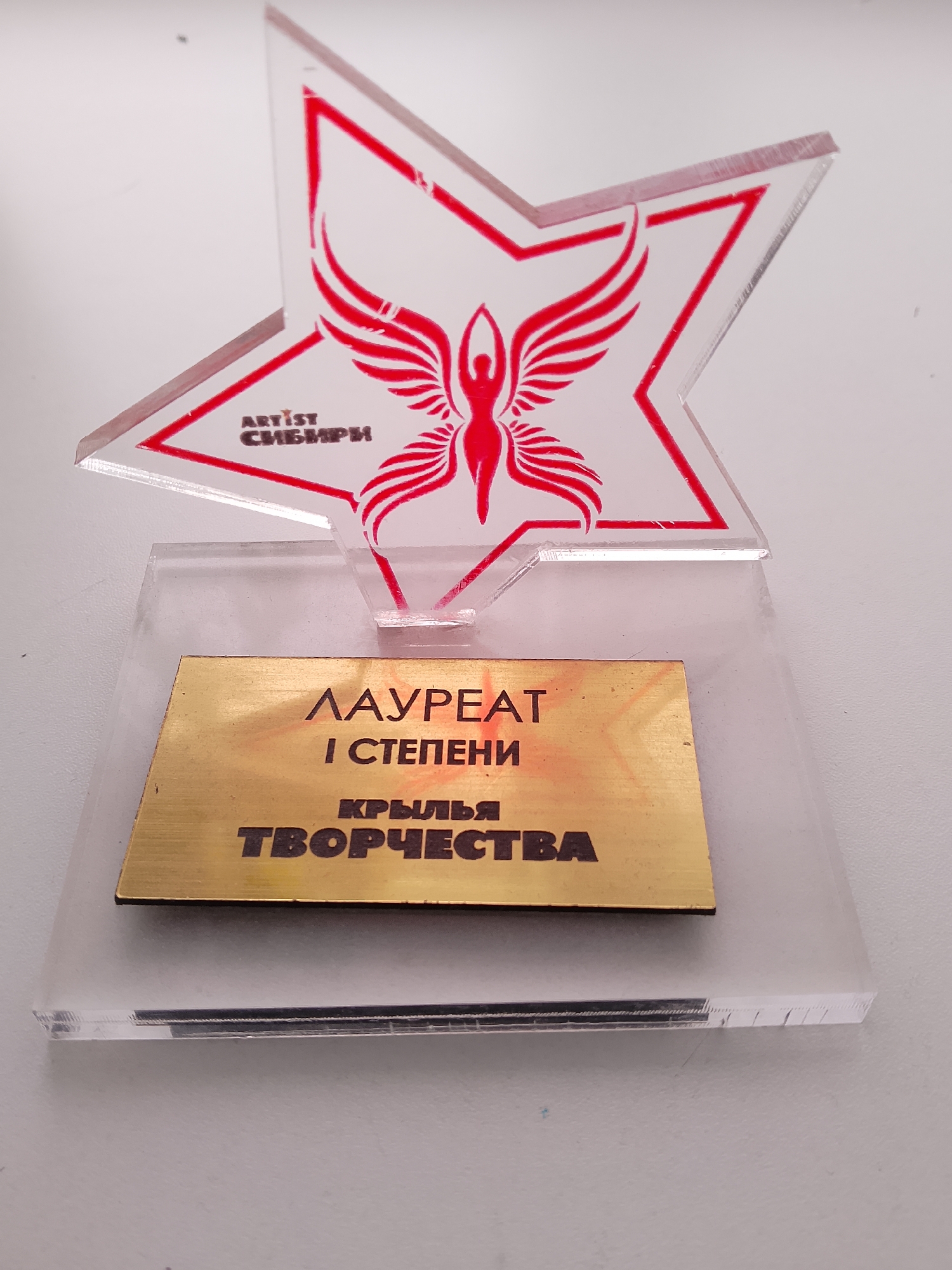 An acrylic award trophy is displayed on a white surface. The trophy features a red graphic of a winged figure within a star-shaped cutout at the top. Below the star, on a gold-colored plaque, is inscribed text in Russian, which translates to "LAUREATE OF THE 1ST DEGREE" and "WINGS OF CREATIVITY". The award is presented on a clear acrylic base. The scene is indoors, with the trophy being the sole subject. No specific location in Abakan, Russia can be determined from the image.