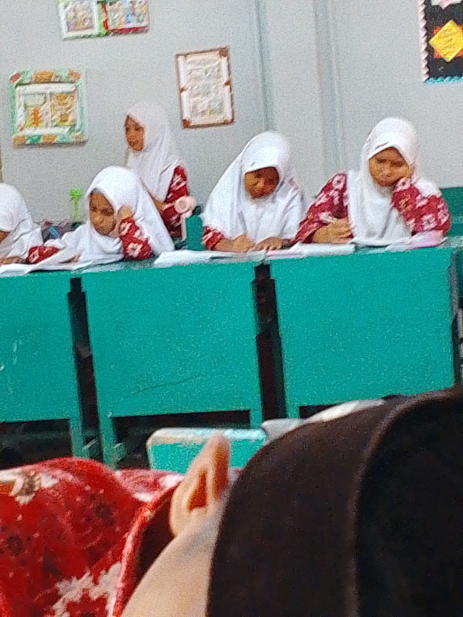 The image captures a classroom setting in Tembung, Indonesia, where several young girls, dressed in white hijabs and patterned blouses, are seated at a teal-colored desk. Two girls in the foreground are actively engaged in writing, their heads bowed over their work. Another girl to their right is resting her chin on her hand, appearing contemplative. In the background, a fourth girl in a hijab stands, looking forward. The classroom walls are adorned with framed pictures. A portion of a person's arm wearing a red and white patterned garment is visible in the lower left corner, and a dark, blurry shape occupies the bottom right of the frame.