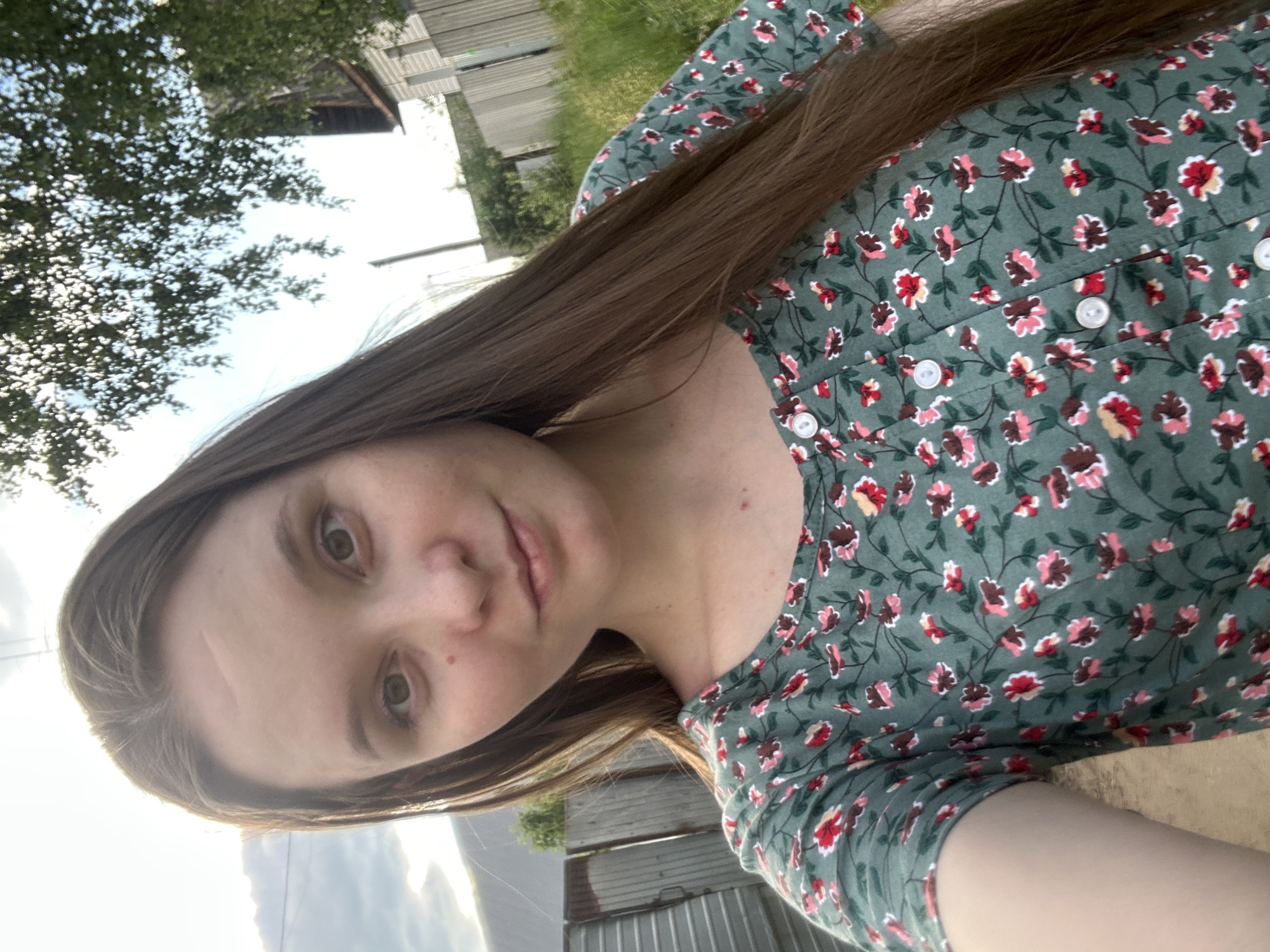 The image depicts a head-and-shoulders selfie of a female subject, estimated to be in her mid-20s, captured outdoors in a rural or suburban residential setting. The subject has long, straight, medium-brown hair, parted on the left and falling over her shoulders. She has a fair complexion, light-colored eyes, and a small mole or freckle located below her right eye (viewer's left). A subtle horizontal crease or scar is visible on her forehead. She is wearing a dark green short-sleeved top patterned with small pink, red, and brown flowers and green foliage, featuring white buttons down the central front placket. She maintains direct eye contact with the camera and displays a faint smile.
The background, stated to be in the city of Chaikovsky, Russia, is illuminated by natural daylight under a partly cloudy sky. To the subject's left, a weathered corrugated metal fence with visible rust runs along what appears to be a dirt or gravel path. Beyond this, a wooden fence and a distant, indistinct building are partially visible. To the subject's right, a substantial tree with green foliage is prominent, partially obscuring the sky. Below the tree, a residential house with light-colored siding and a dark roof, a utility pole, and various fences bordering grassy areas are present.