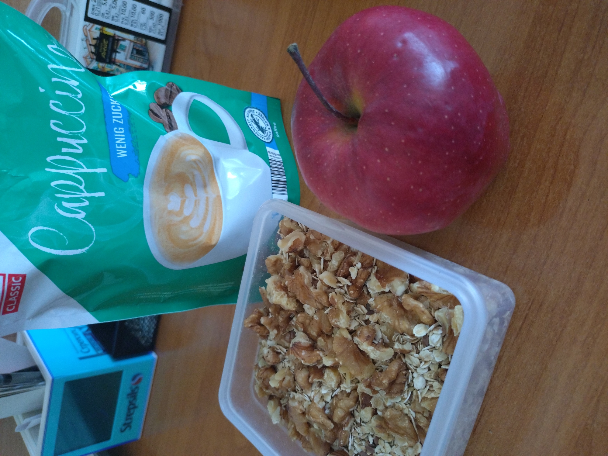 The image shows a tabletop still life with a focus on food items. The items are arranged on a wooden surface. A package of "Cappuccino Wenig Zucker" is lying on the left side of the image. Next to it is a blue box labeled "Strepsils". To the right of the cappuccino package is a red apple. A clear plastic container filled with walnuts and oats is positioned at the bottom center of the frame. The OCR data indicates text on the objects, and includes pricing information and the phrase "TOPLE & NATURE Kaffee" on the cappuccino package. The image gives the impression of a breakfast or snack setting.